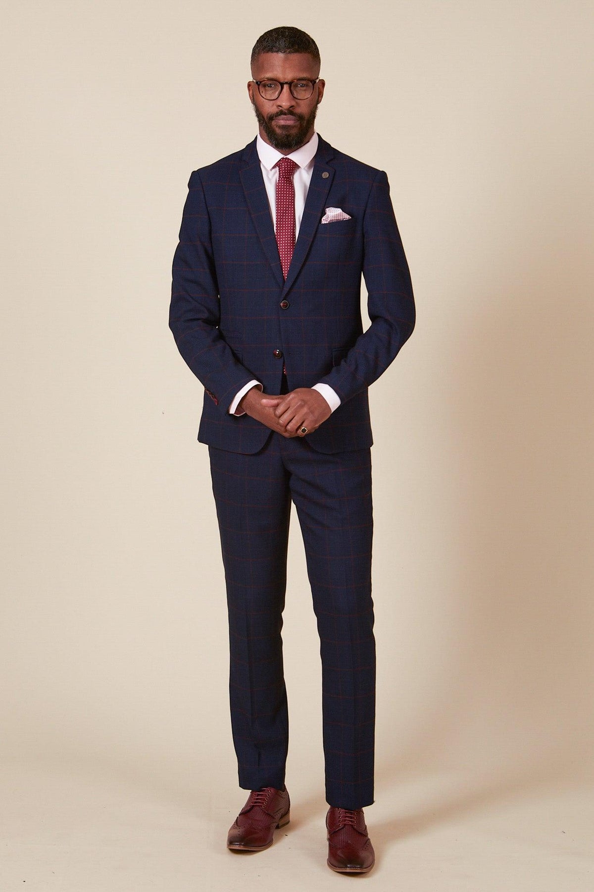 A man stands against a plain background wearing the Marc Darcy EDINSON Navy Wine Check Two Piece Suit with a slim fit blazer, white dress shirt, red patterned tie, and burgundy shoes. He has short hair, a beard, glasses, and his hands are clasped in front.