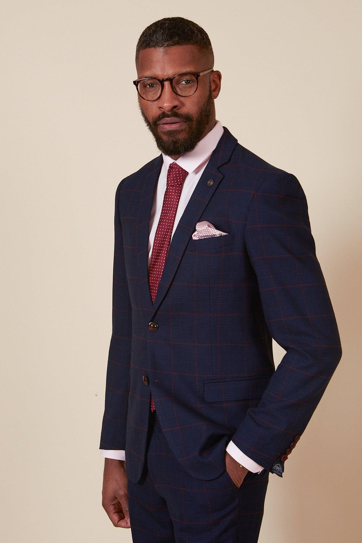 A man with short hair and a beard wears glasses, the Marc Darcy EDINSON Navy Wine Check Two Piece Suit, a white shirt, patterned red tie, and pink pocket square against a plain light background.