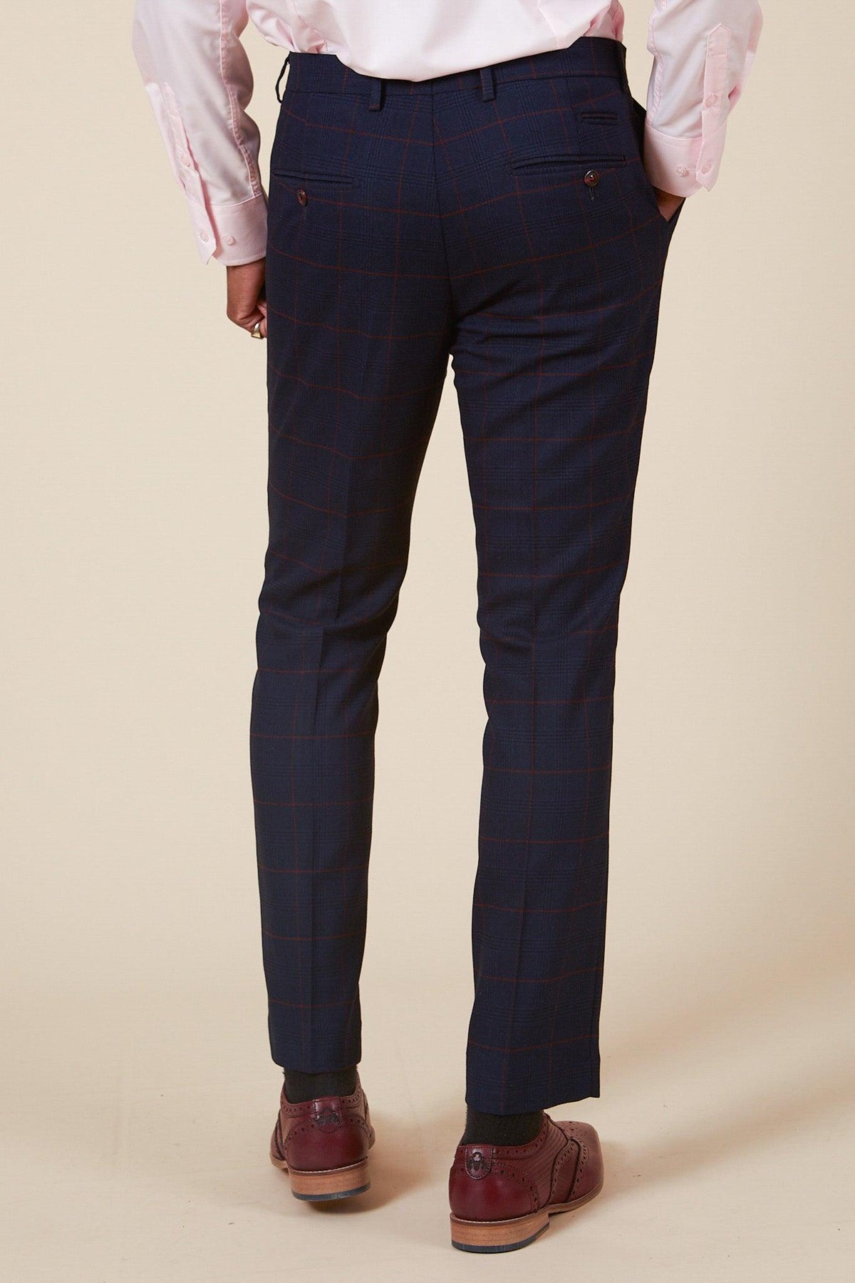A person stands facing away with hands in their pockets, wearing the Marc Darcy EDINSON Navy Wine Check Two Piece Suit and a light pink dress shirt, paired with brown dress shoes against a plain beige background.