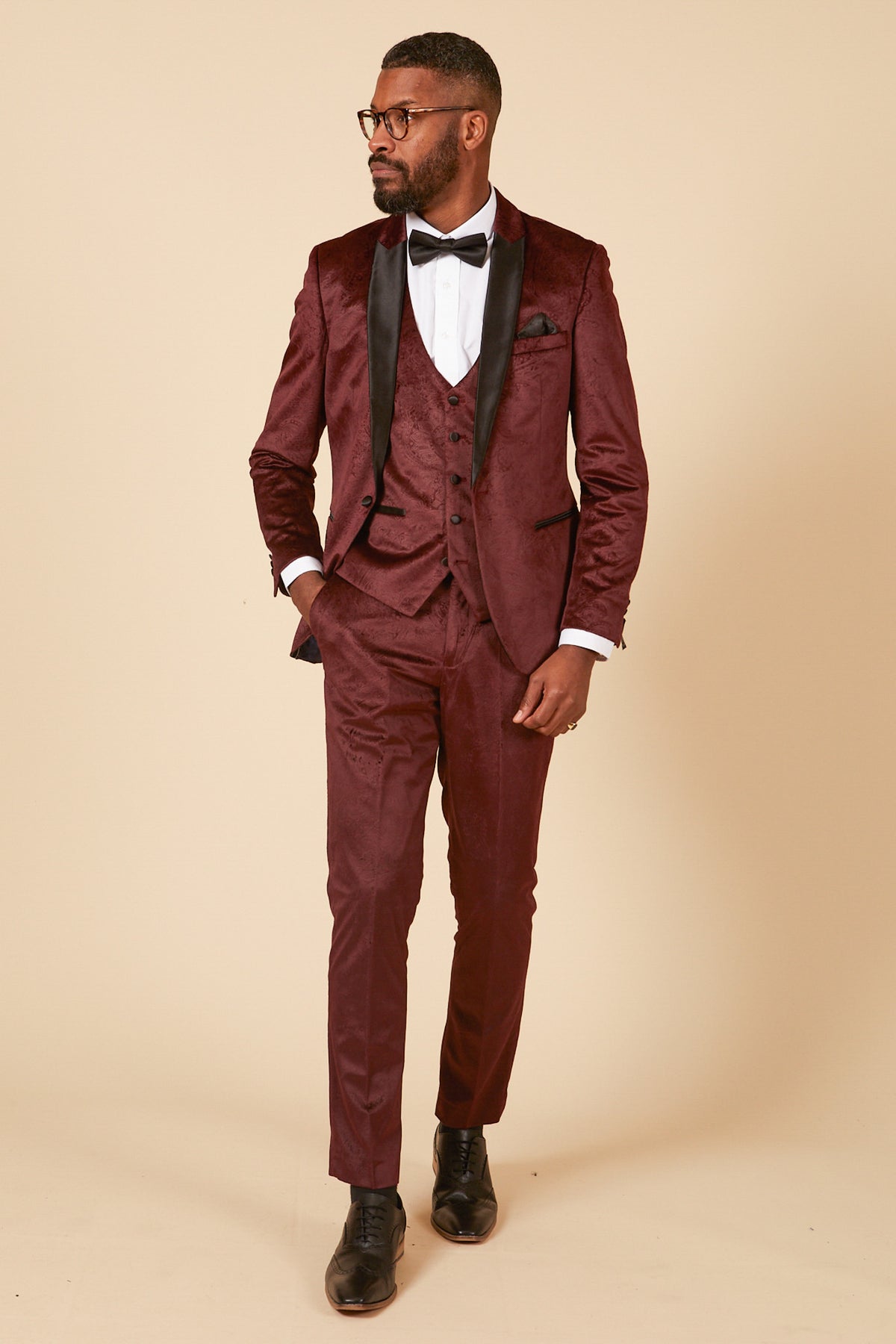 A man in the Marc Darcy SIMON wine velvet jacquard three piece suit, styled with a black satin lapel, white shirt, black bow tie, and dress shoes, stands against a beige background—showcasing refined party elegance with short hair, beard, and glasses.