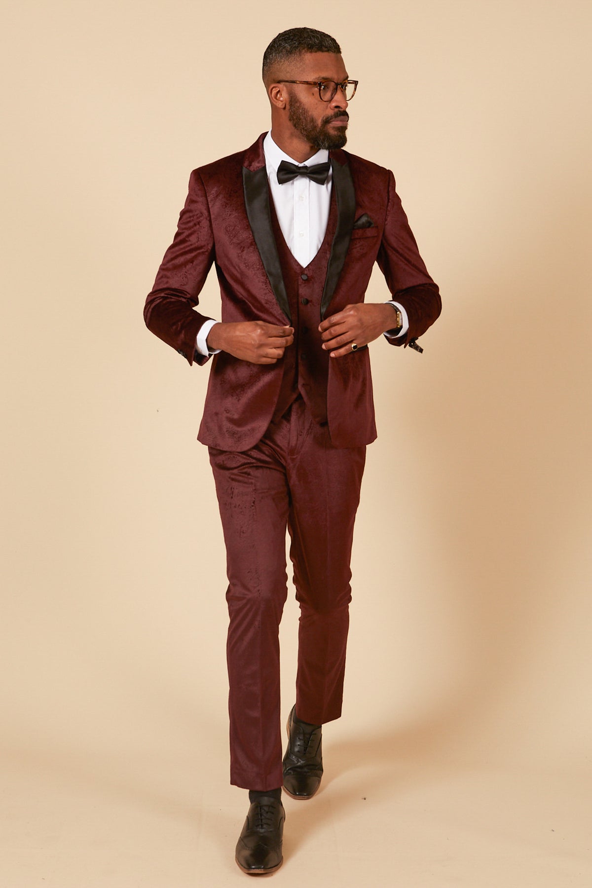 A man showcases the Marc Darcy SIMON Wine Velvet Jacquard Three Piece Suit, featuring sleek black lapels. Against a beige backdrop, he adjusts his jacket and glances sideways, embodying sophisticated party wear style.