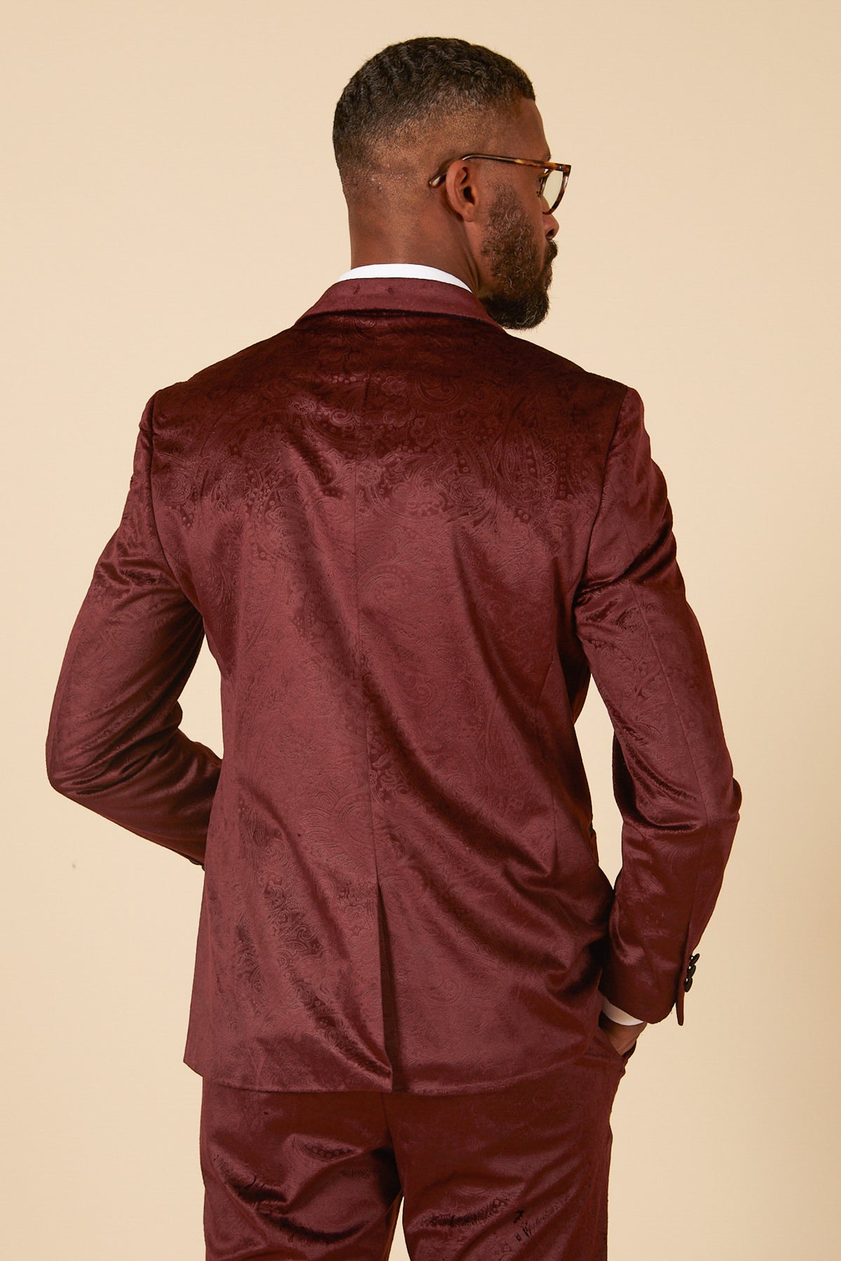 A man with short hair, a beard, and glasses wears the Marc Darcy SIMON Wine Velvet Jacquard Three Piece Suit, standing with one hand in his pocket against a beige background—ideal for standout party style.