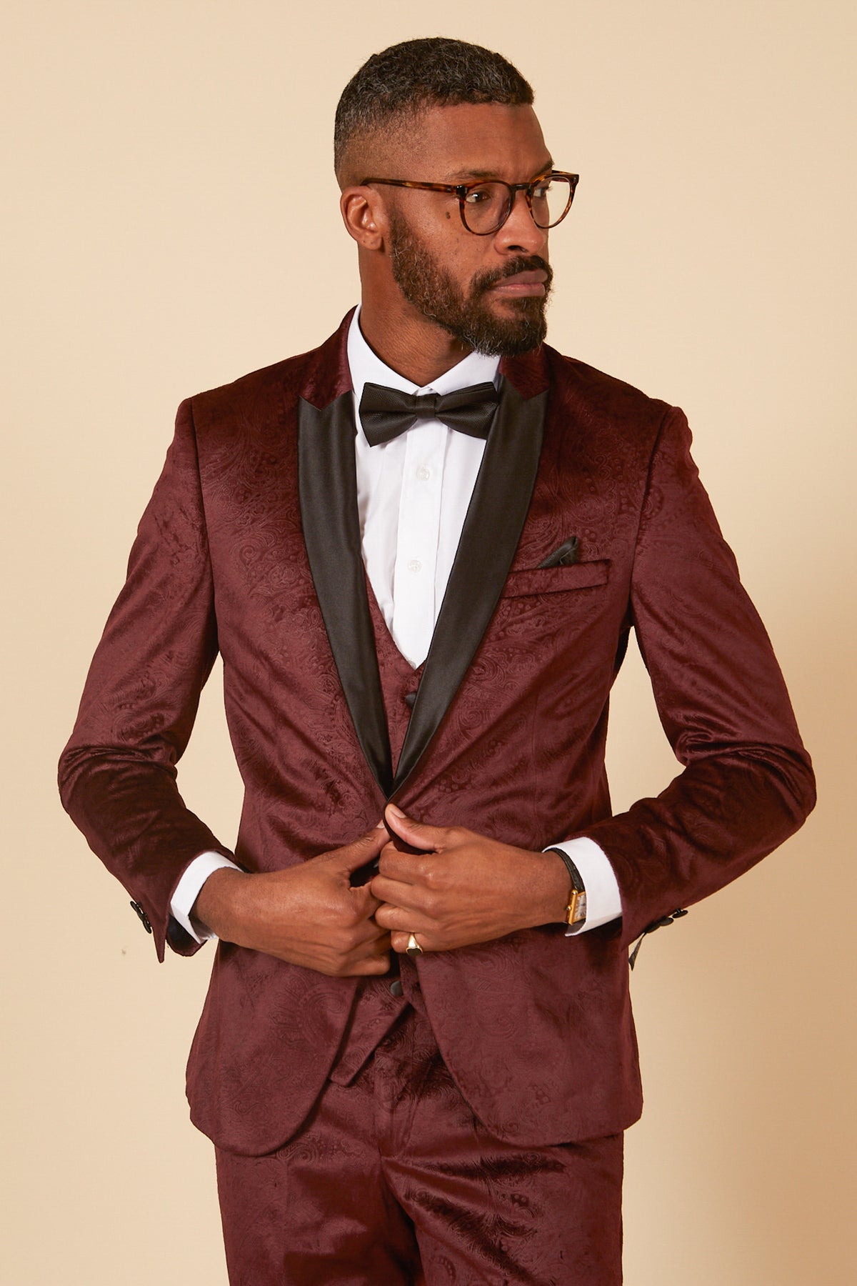 A man in the Marc Darcy SIMON Wine Velvet Jacquard Three Piece Suit stands against a beige background, buttoning his jacket and looking to the side—a sophisticated choice for elegant party occasions.