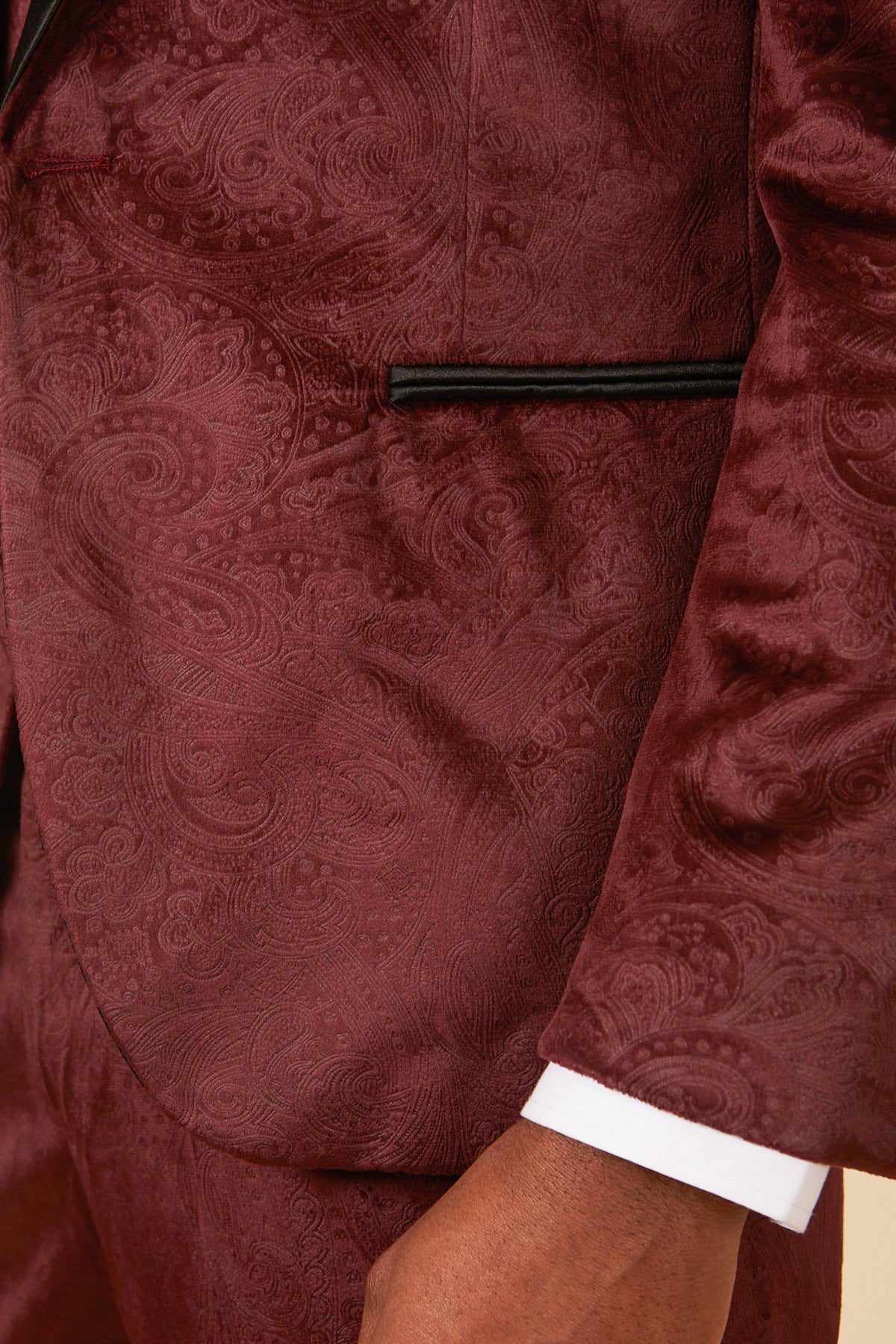 Close-up of a person wearing the Marc Darcy SIMON Wine Velvet Jacquard Three Piece Suit—ideal for parties. The photo shows a maroon paisley-patterned jacket, black-trimmed pocket, visible hand, and white shirt cuff.