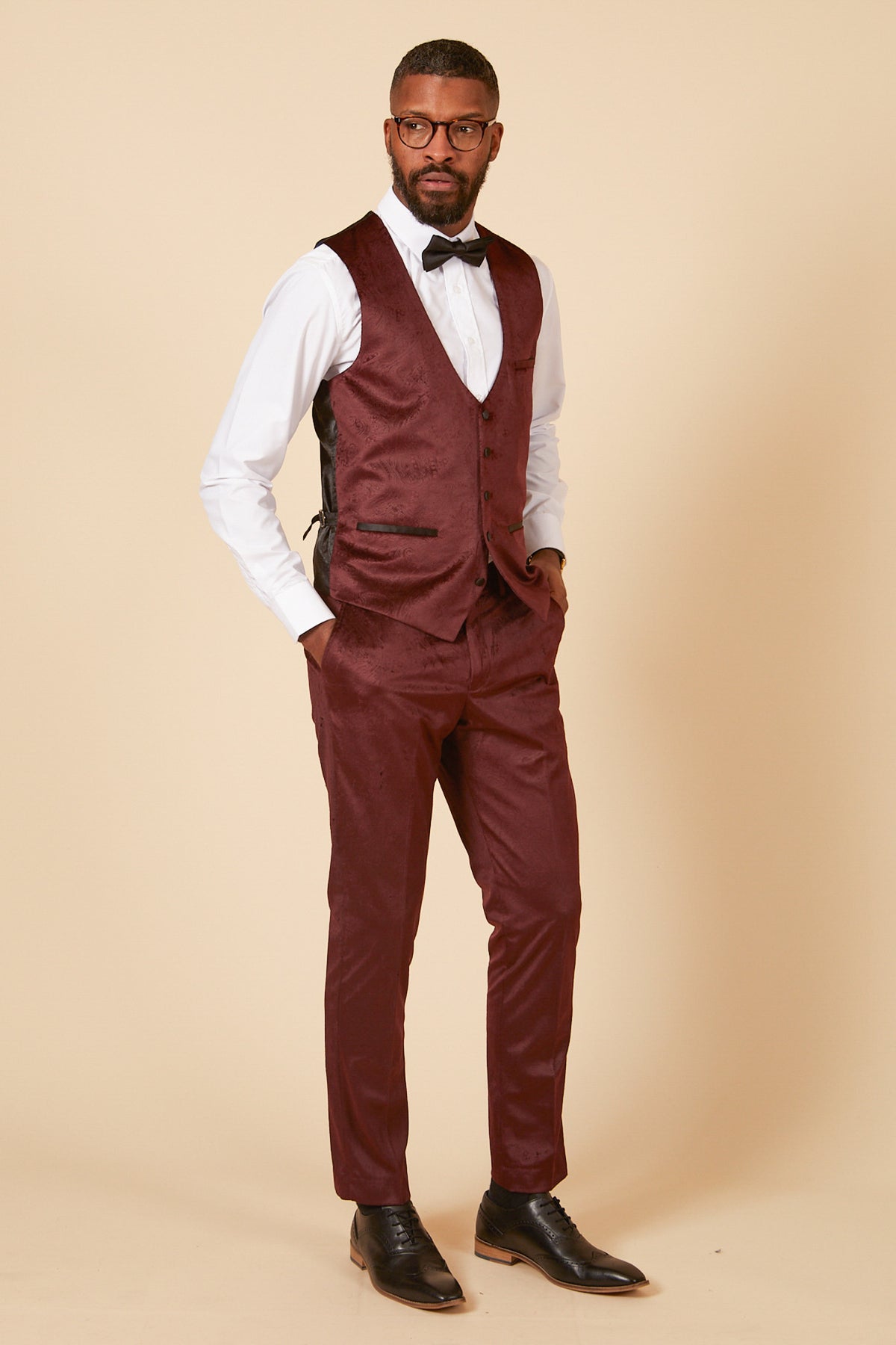 A man wears the Marc Darcy SIMON Wine Velvet Jacquard Three Piece Suit—ideal for parties—with a white shirt, black bow tie, glasses, and dress shoes. He stands against a beige background with hands in pockets, gazing to the side.