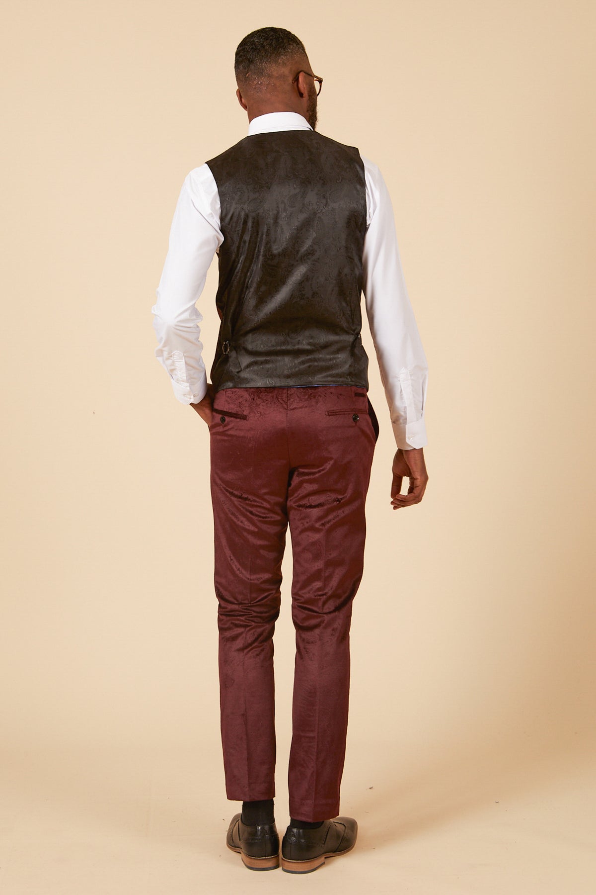 A man stands with his left hand in his pocket, wearing the Marc Darcy SIMON Wine Velvet Jacquard Three Piece Suit, white shirt, and dark shoes against a beige background.
