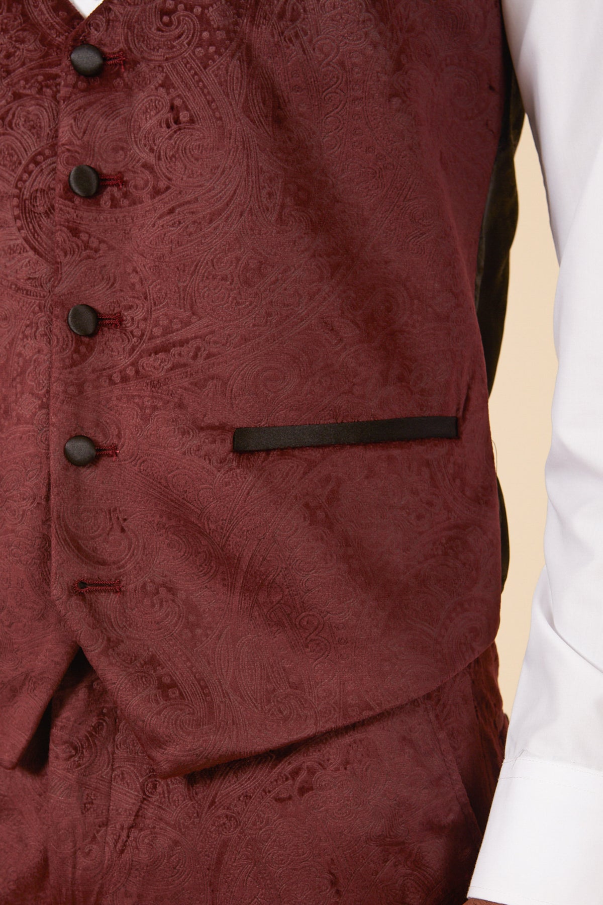 Close-up of a person wearing the Marc Darcy SIMON - Wine Velvet Jacquard Three Piece Suit, featuring a detailed paisley pattern, black buttons, and black-trimmed pocket over a white dress shirt. Only the torso and partial arm are visible.