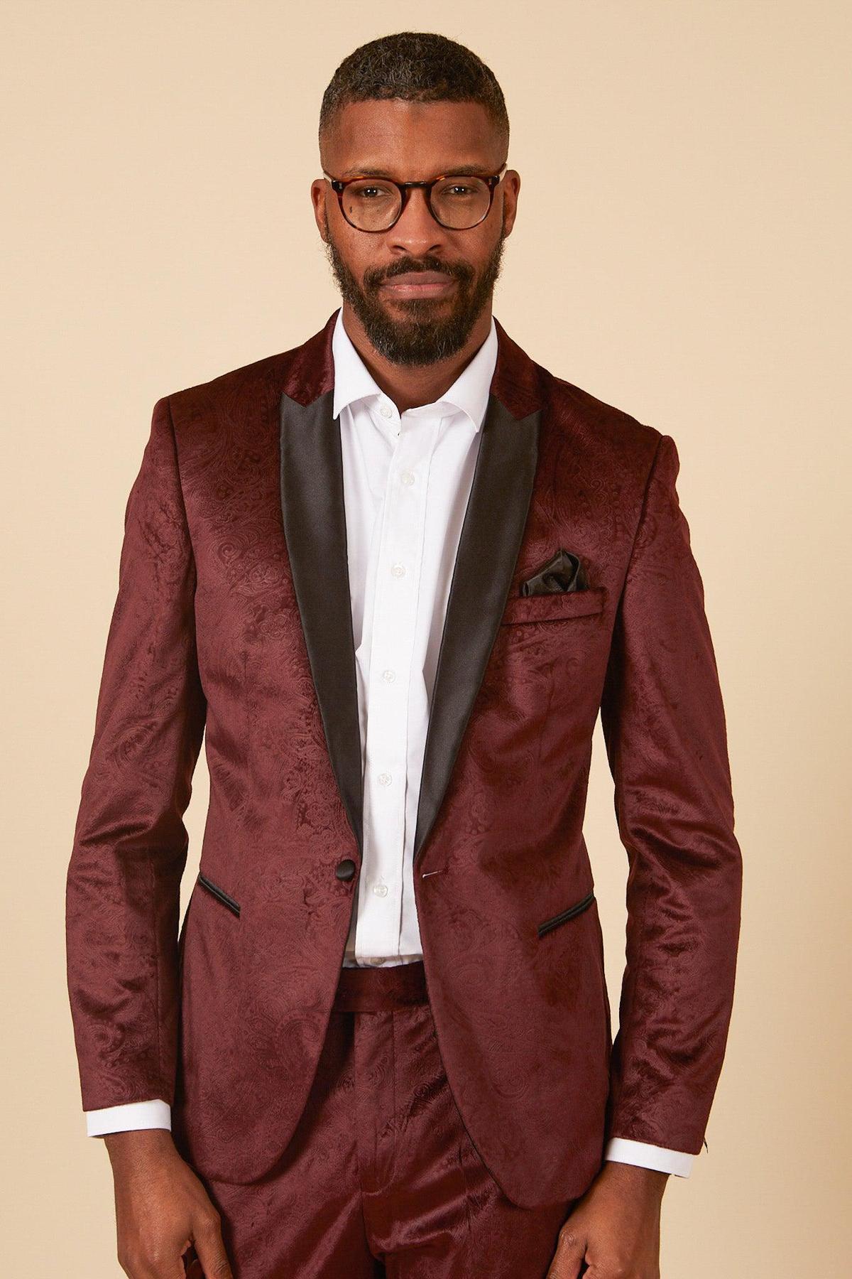 A man with short hair, a beard, and glasses wears a white shirt and the marcdarcy SIMON - Velvet Wine Tux Lapel Jacquard Blazer, featuring black lapels and a subtle paisley pattern, standing against a plain beige background.