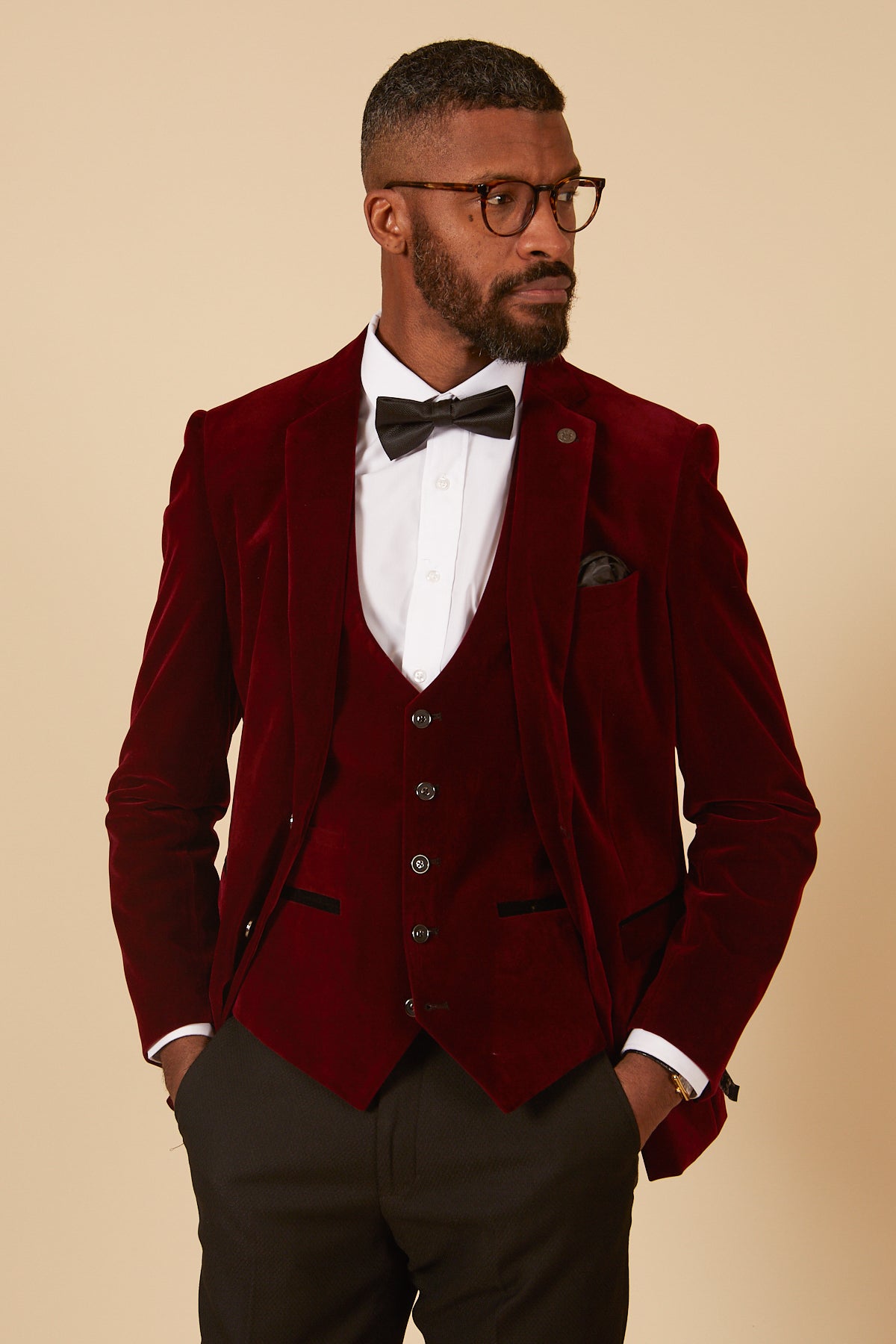 A man in glasses, a white dress shirt, black bow tie, and the marcdarcy HUDSON Wine Velvet Blazer with matching vest stands with hands in pockets, looking to the side against a beige background.