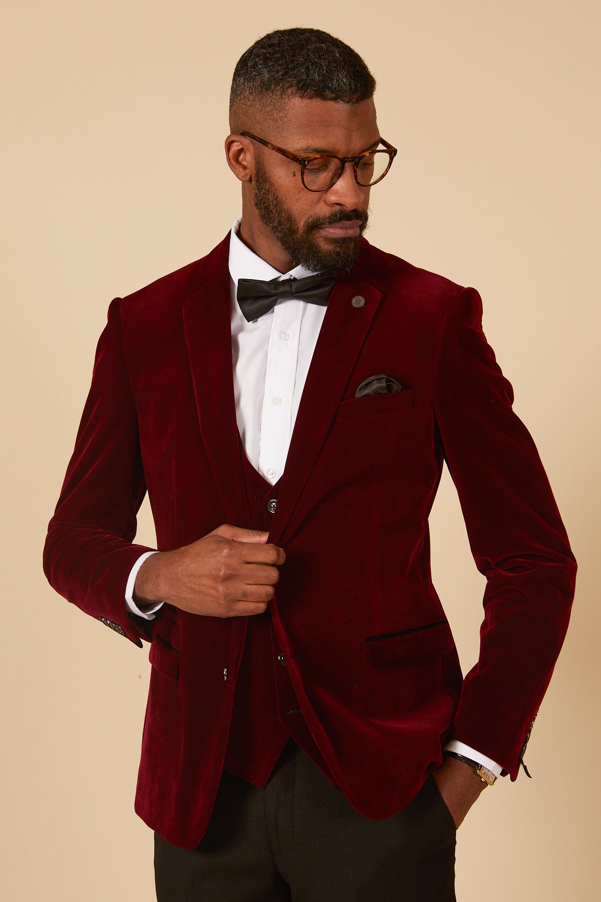 A man in glasses, a white dress shirt, black bow tie, and the marcdarcy HUDSON - Wine Velvet Blazer with a pocket square stands against a beige background, looking down and adjusting his jacket.