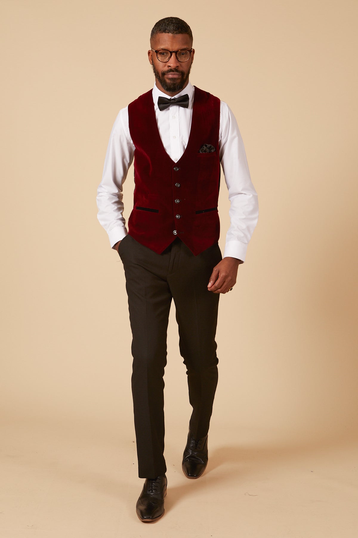 A man in glasses, a white dress shirt, black bow tie and pants walks confidently against a beige background wearing the marcdarcy HUDSON Wine Velvet Jacquard Waistcoat in a tailored fit.