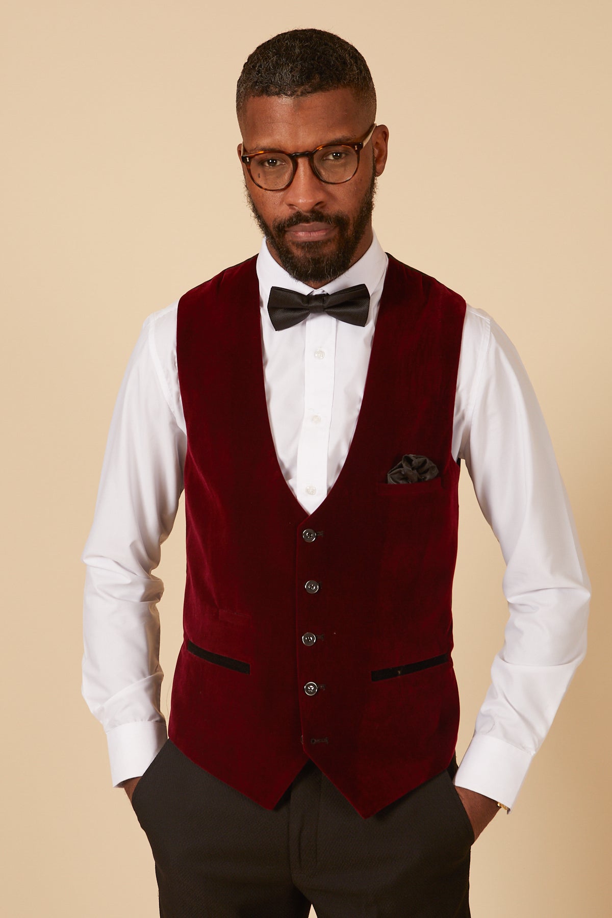 A Black man wears a marcdarcy HUDSON Wine Velvet Jacquard Waistcoat in a tailored fit, paired with a white dress shirt, black bow tie, and glasses. His hands rest in his vest pockets.