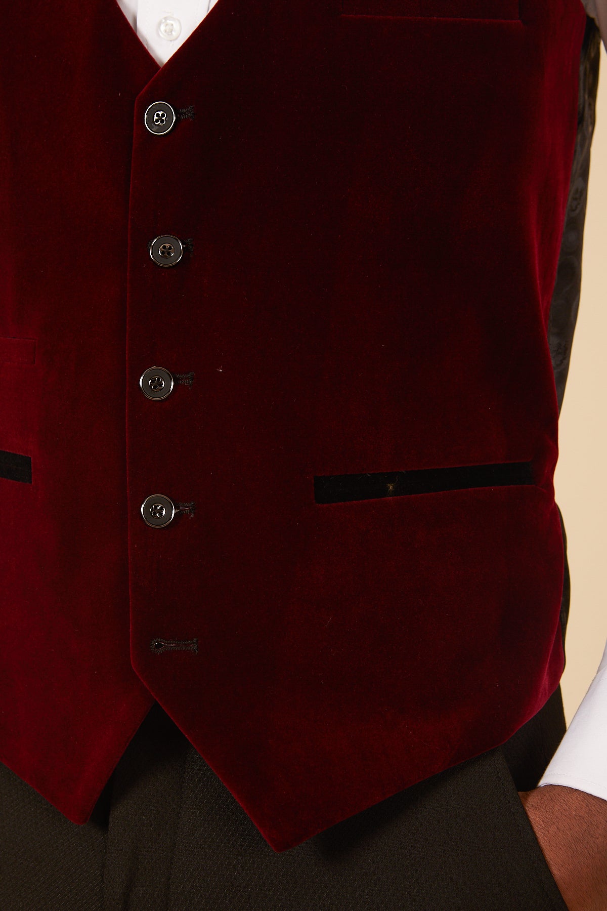 A close-up of a person wearing the marcdarcy HUDSON Wine Velvet Jacquard Waistcoat in a tailored fit with black buttons, two black-trimmed pockets, and a sharply pointed hem over a white shirt and black trousers.