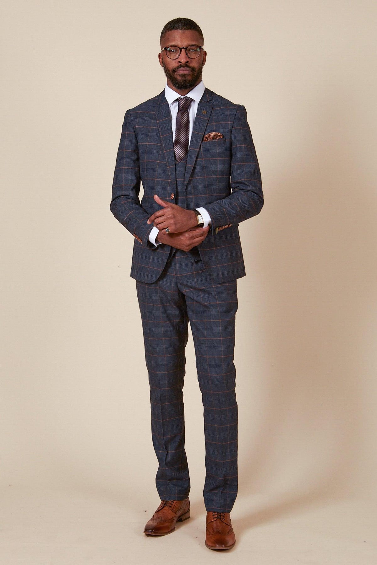A man stands against a beige background wearing marcdarcy’s The Everton Collection—JENSON Marine Navy Check Suit, paired with a white shirt, brown tie and shoes, glasses, and a pocket square, confidently looking at the camera with hands clasped.