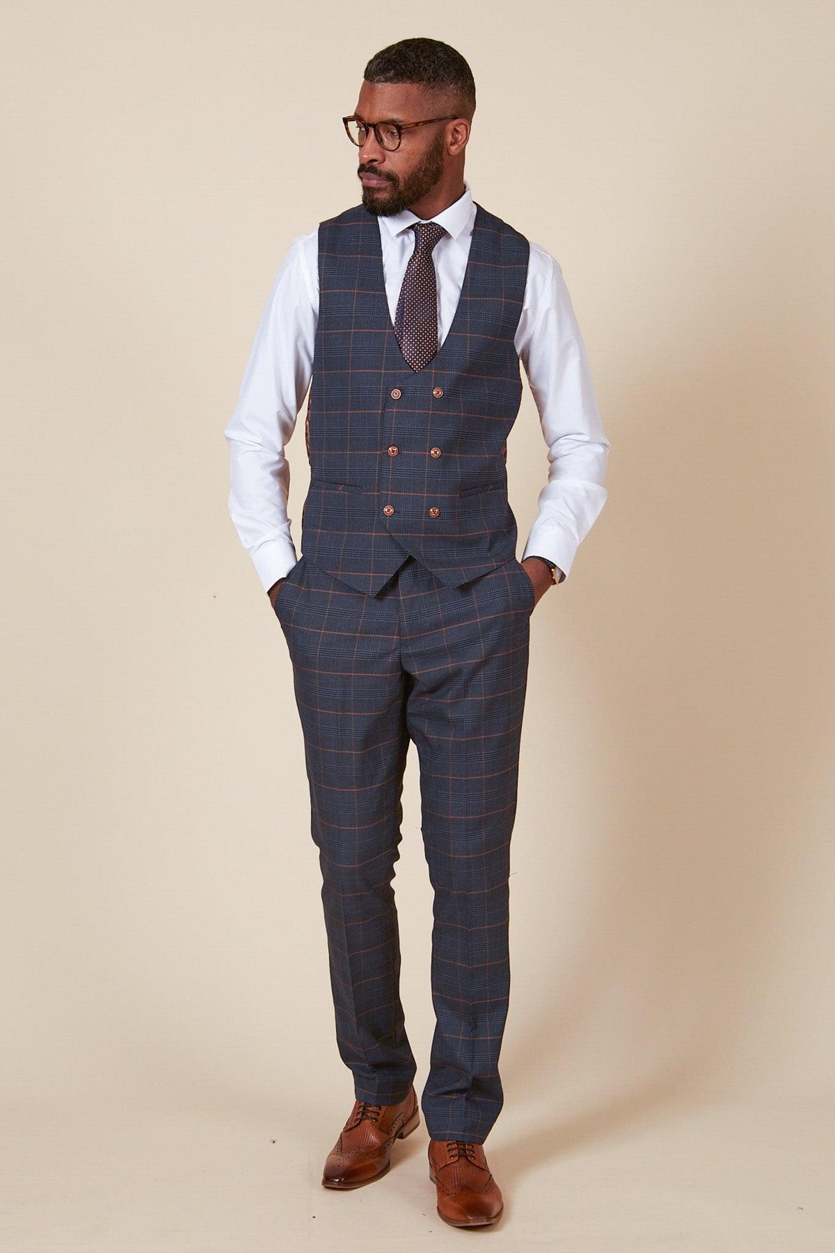 A man wearing glasses, a white shirt, the marcdarcy JENSON - Marine Navy Check Double Breasted Waistcoat with matching trousers, a patterned tie, and brown dress shoes stands with hands in pockets against a beige background.