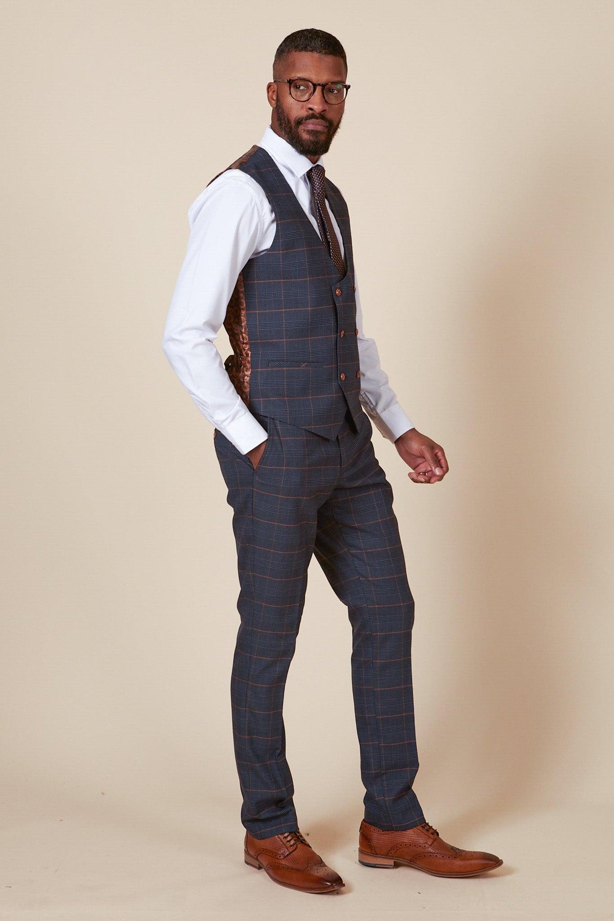 A man with short hair and a beard wears glasses, a white shirt, striped tie, brown shoes, and the marcdarcy JENSON - Marine Navy Check Double Breasted Waistcoat. He stands sideways with one hand in his pocket against a plain light background.