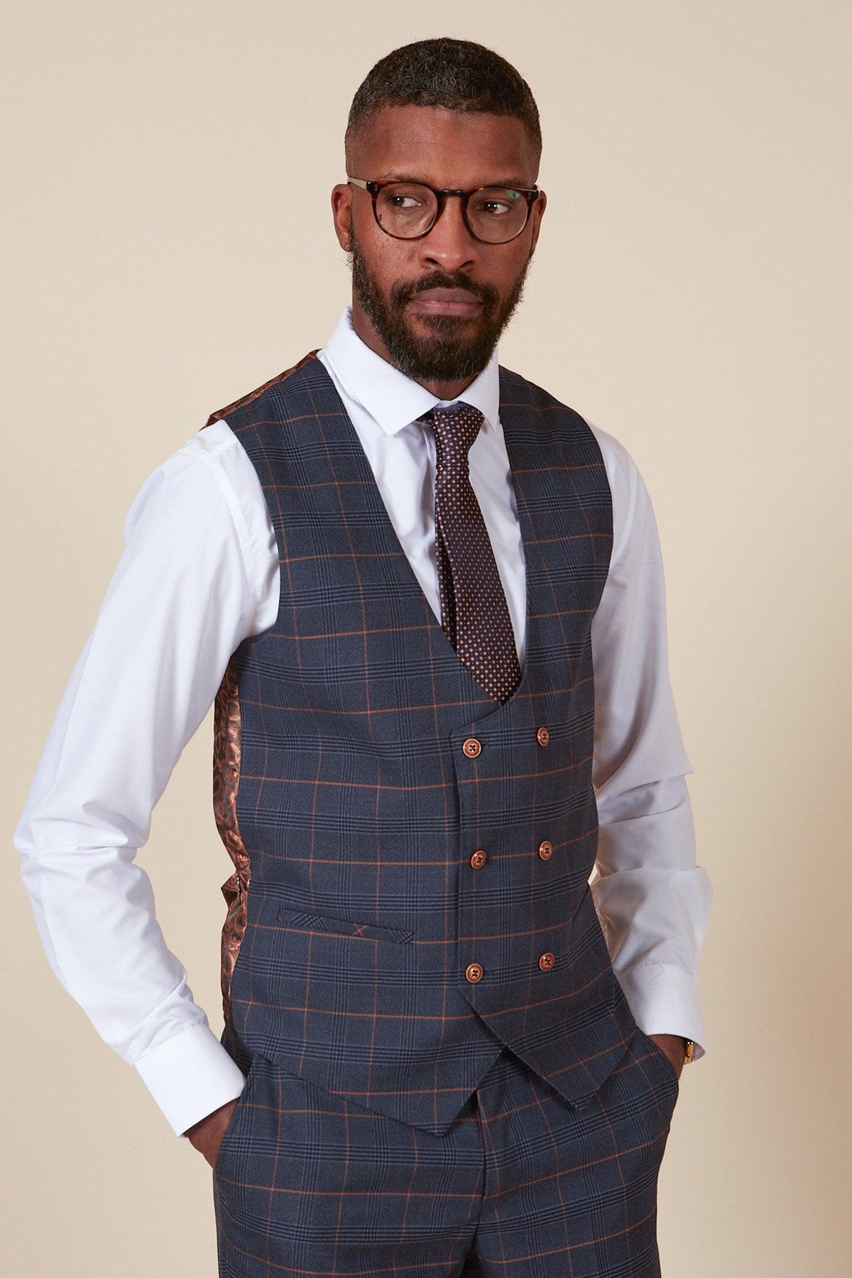 A man wearing glasses, a white dress shirt, a patterned tie, and the marcdarcy JENSON - Marine Navy Check Double Breasted Waistcoat with matching slim fit trousers stands against a beige background, looking slightly to the side.