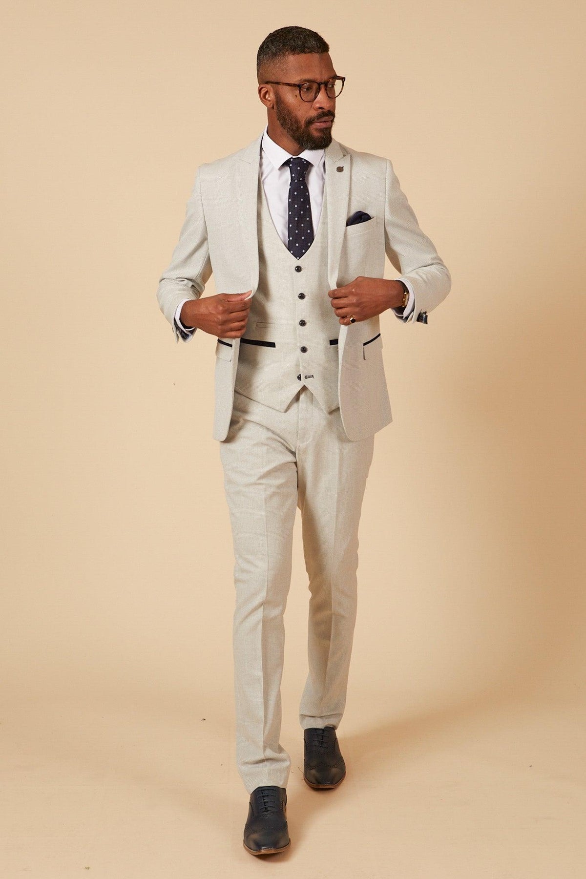 Former Manchester City footballer Fabian Delph wears the Marc Darcy BROMLEY Stone Check Suit, paired with a white shirt, navy polka dot tie, navy pocket square, and dark dress shoes, standing against a beige background.