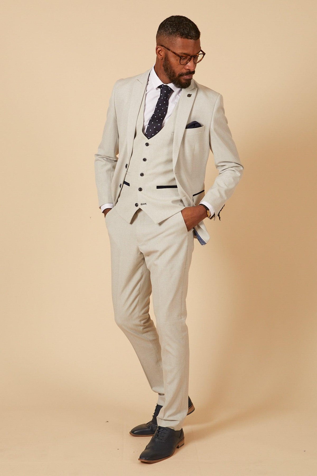 Former Manchester City footballer Fabian Delph wears a Marc Darcy BROMLEY Stone Check Suit, posing against a beige background with one hand in his pocket, looking down and to the side.