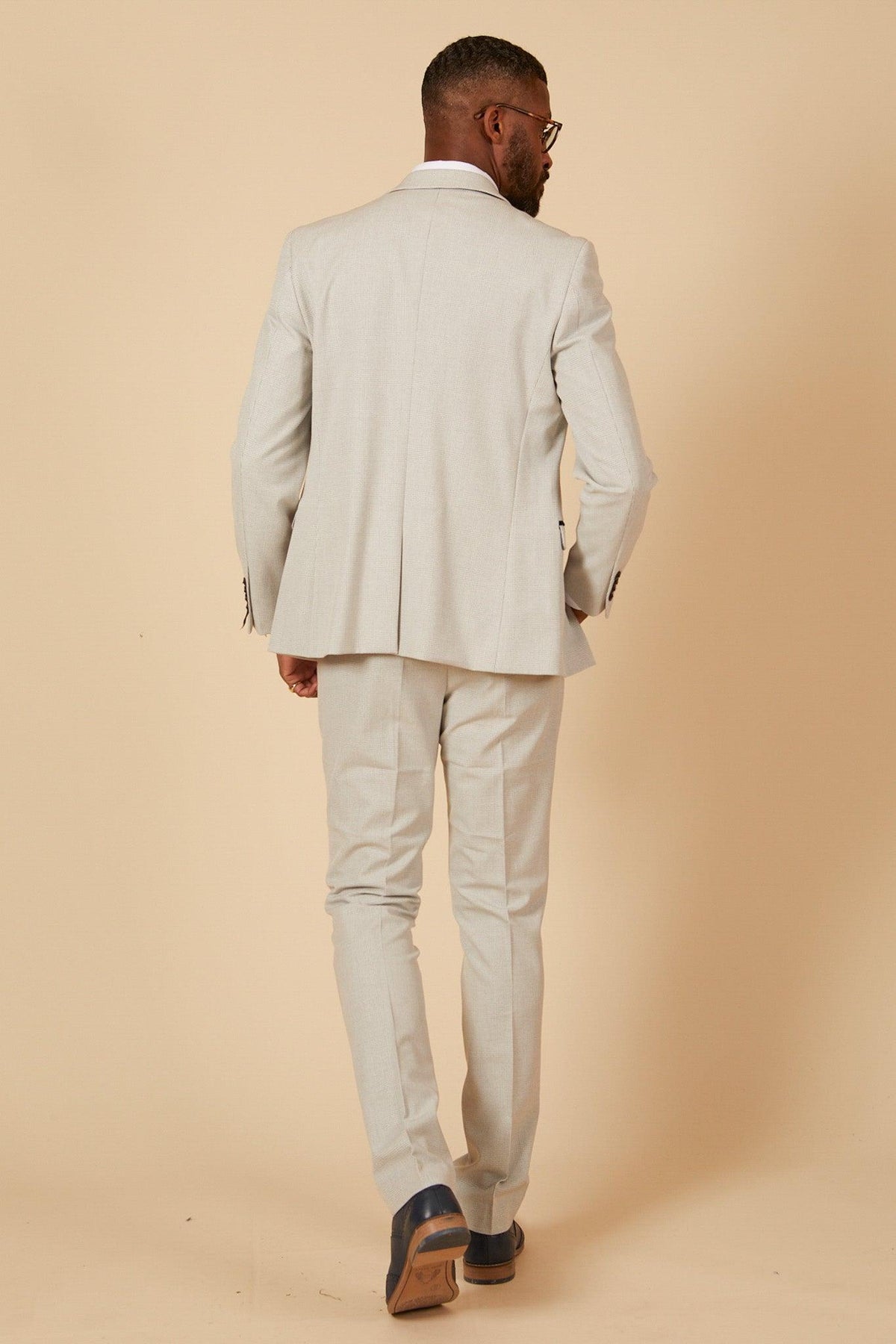 Former Manchester City footballer Fabian Delph is seen from behind in a Marc Darcy BROMLEY Stone Check Suit, exuding refined style against a beige background. He sports short hair, glasses, and brown dress shoes.