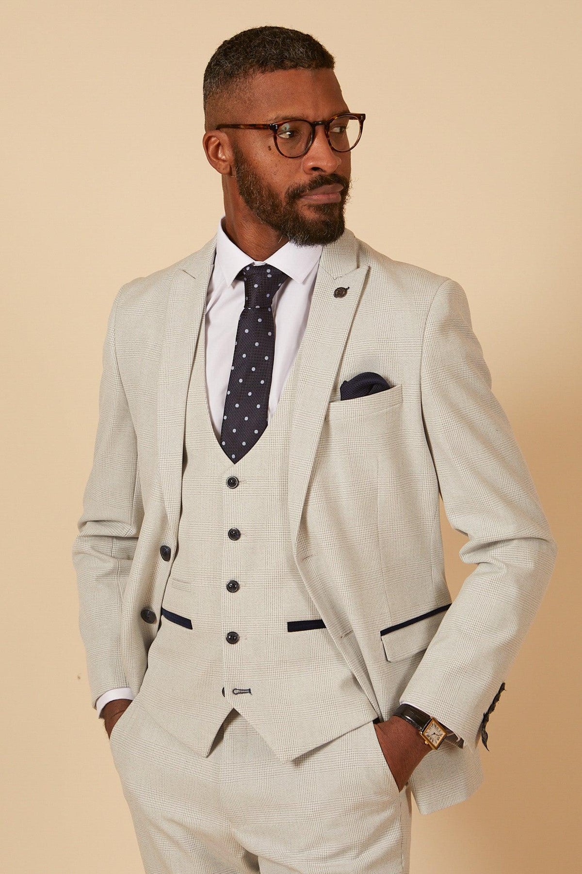 Former Manchester City footballer Fabian Delph wears the Marc Darcy BROMLEY Stone Check Suit, styled with a navy polka-dot tie, dark blue pocket square, glasses, and a watch.