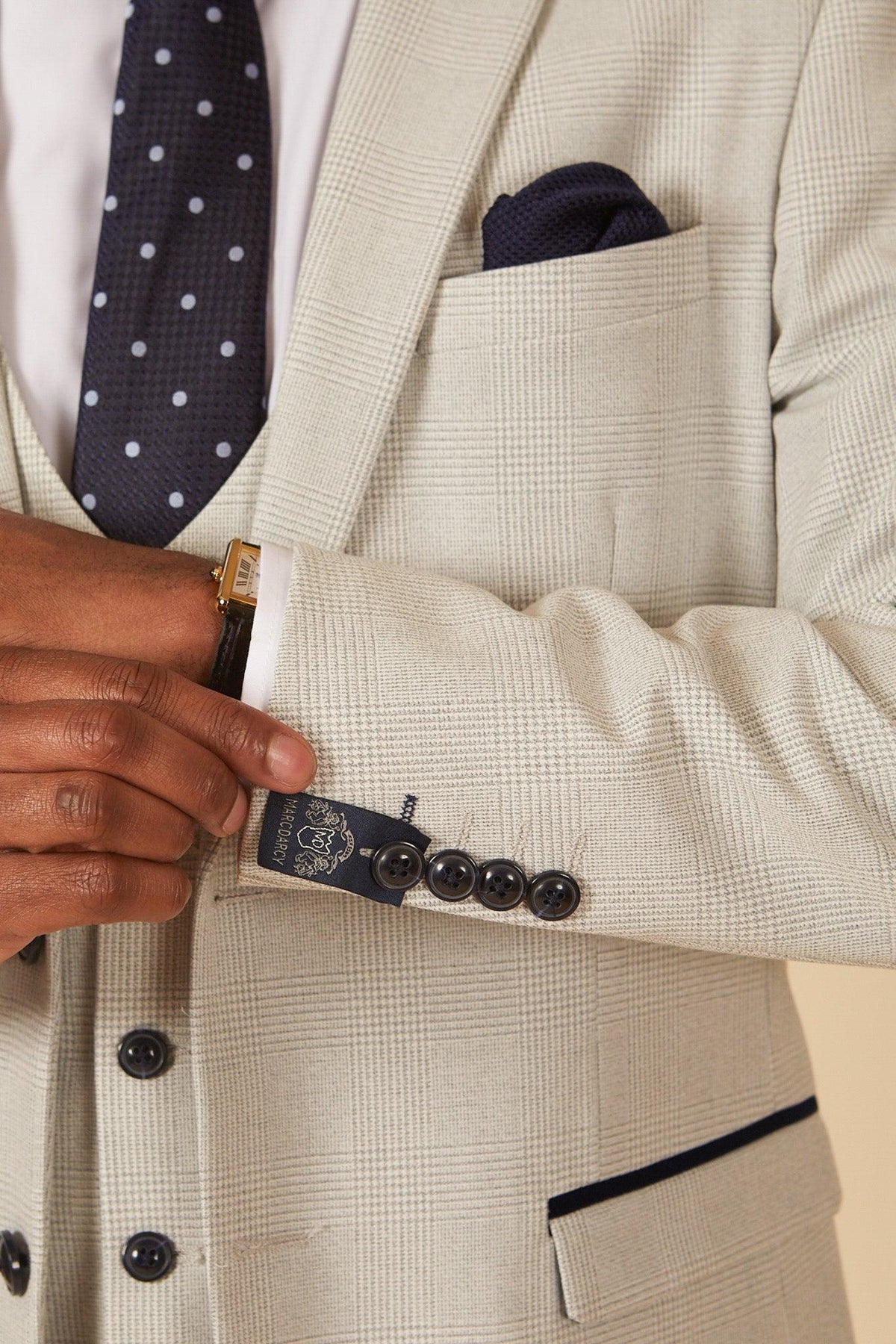Former Manchester City footballer Fabian Delph wears the Marc Darcy BROMLEY Stone Check Suit, featuring a slim fit, dark buttons, white shirt, navy polka dot tie, and matching pocket square. Only his torso and arm are visible as he adjusts his cuff.