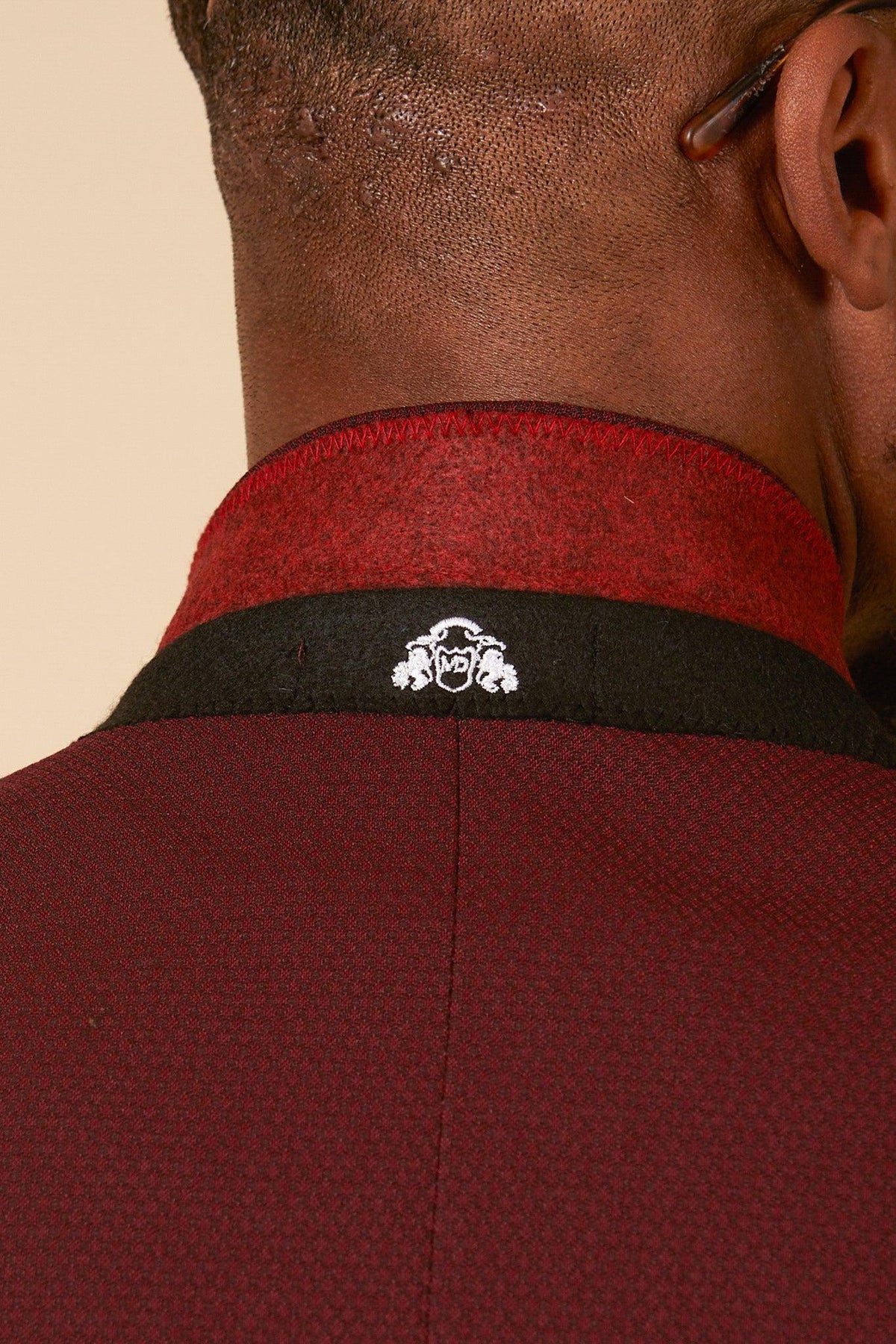 Close-up of the back of a persons neck wearing the marcdarcy MAX - Wine Three Piece Suit. The black and red collar features a small white embroidered emblem. The persons ear and short hair are partially visible.