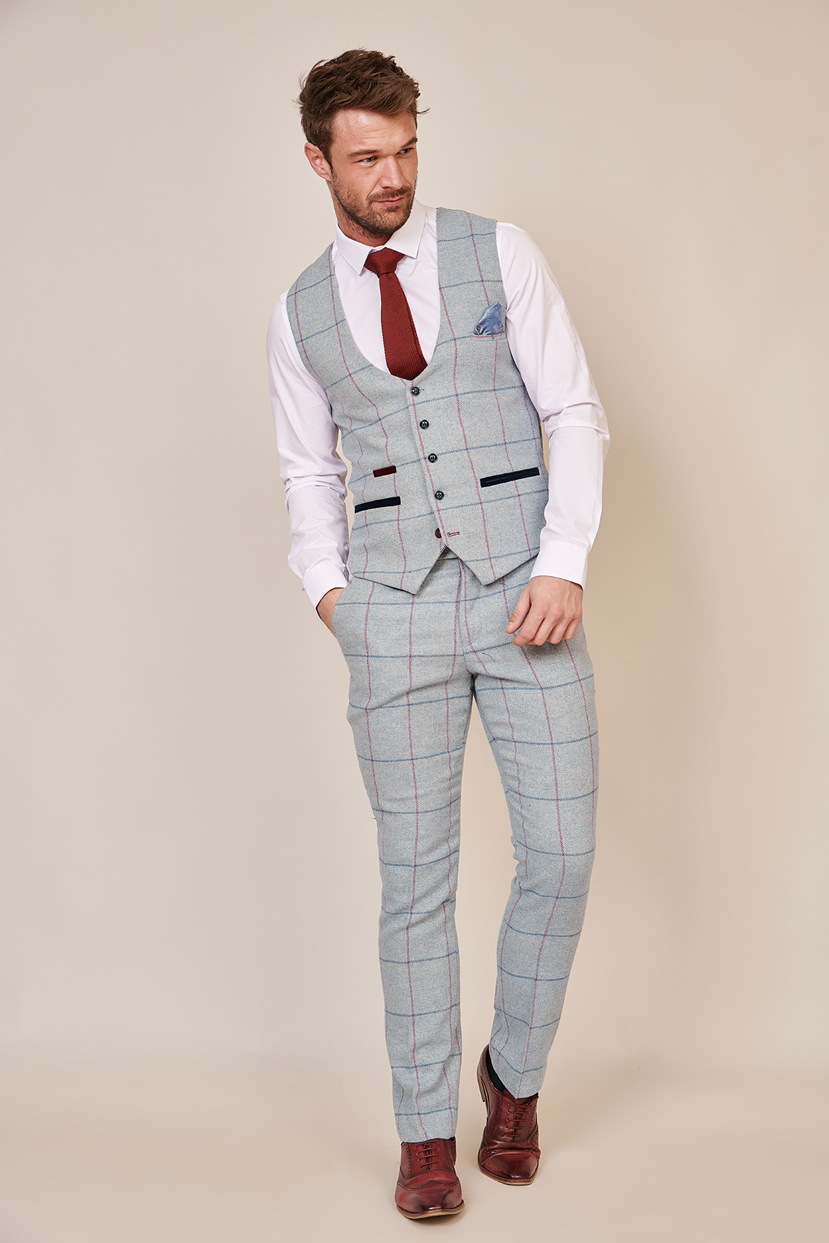 A man wearing the Marc Darcy NICOLAS - Sky Tweed Check Waistcoat with a white shirt, red tie, and brown shoes stands against a beige background, looking down and slightly to the side.