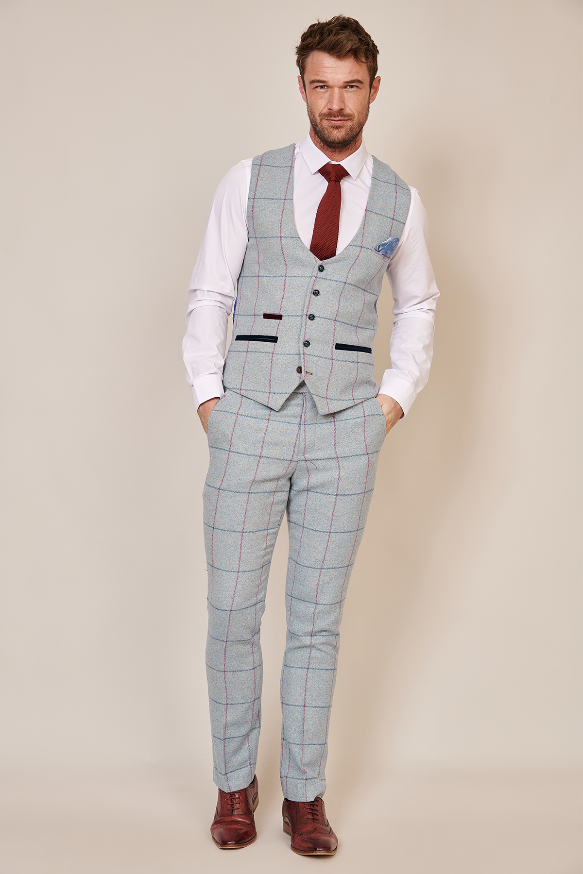 A bearded man stands with hands in pockets wearing the Marc Darcy NICOLAS Sky Tweed Check Waistcoat over a white shirt, paired with a light gray plaid suit, red tie, and brown dress shoes against a plain beige background.