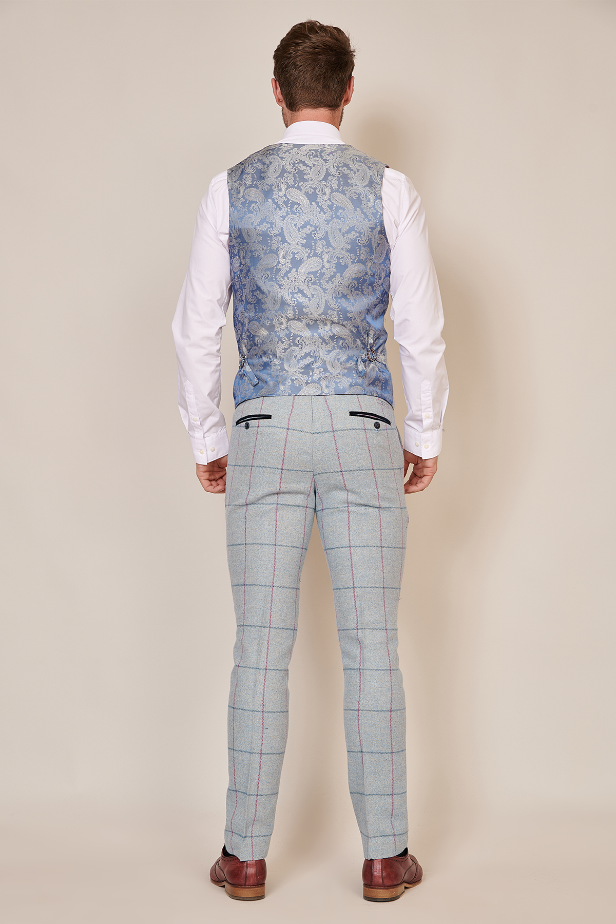 A man faces away from the camera, wearing a Marc Darcy NICOLAS - Sky Tweed Check Waistcoat, a white dress shirt, light blue plaid trousers with a pink grid, and brown dress shoes.