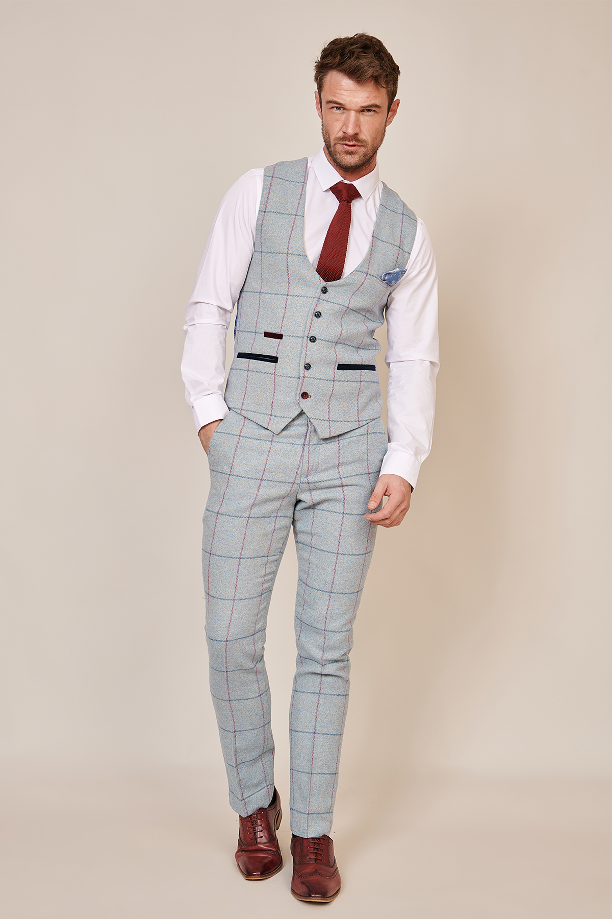 A man wearing the Marc Darcy NICOLAS Sky Tweed Check Waistcoat with a light gray plaid suit, white shirt, red tie, and brown shoes stands against a plain beige background with one hand in his pocket.