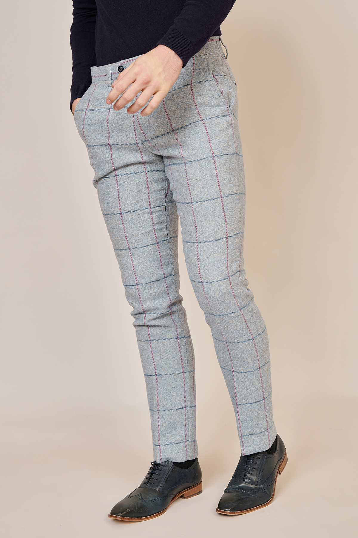 Wearing Marc Darcy’s NICOLAS - Sky Check Tweed Trousers, a slim fit style in light gray plaid with red and blue lines, plus a dark long-sleeve shirt and black dress shoes, a person stands hands in pockets against a plain background.