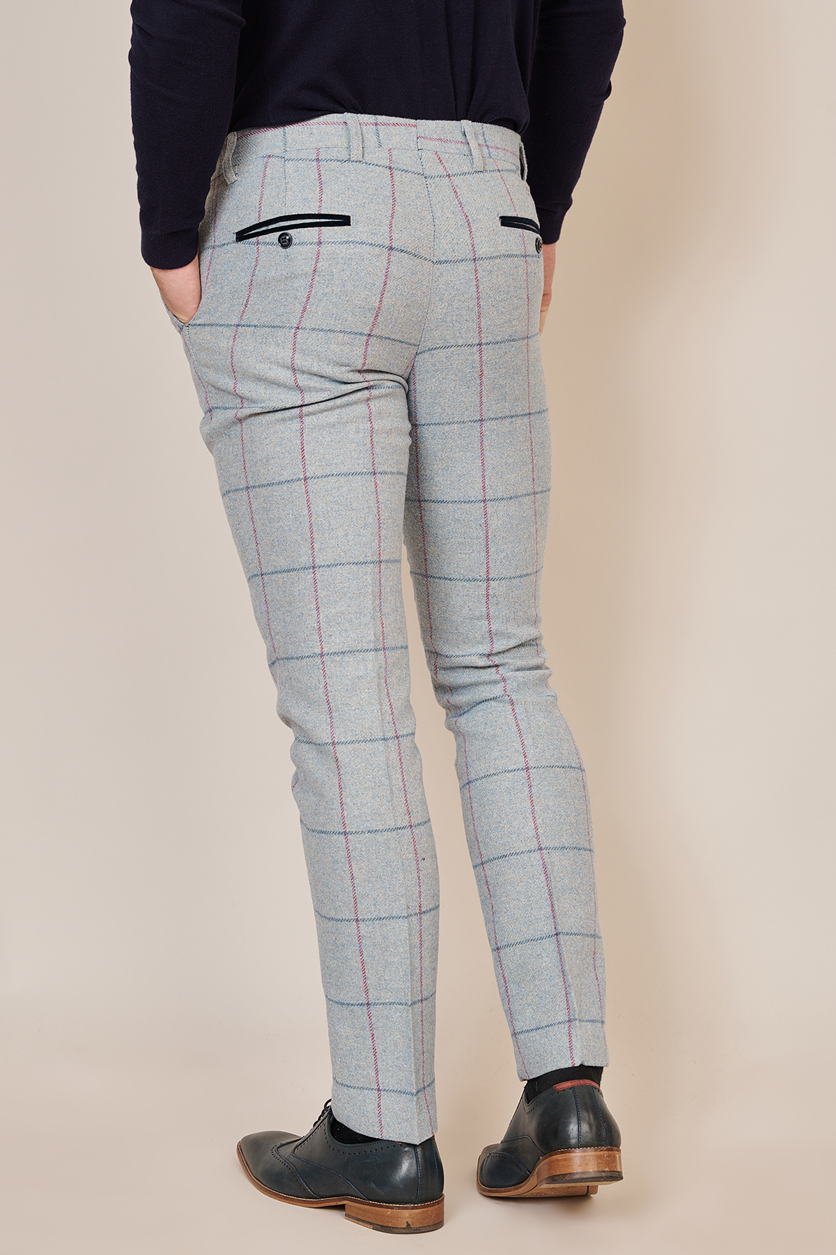 A person faces away wearing Marc Darcy's NICOLAS - Sky Check Tweed Trousers, featuring a slim fit and light gray plaid with red and blue lines, paired with a black top and black dress shoes. Hands in pockets reveal the stylish back view.