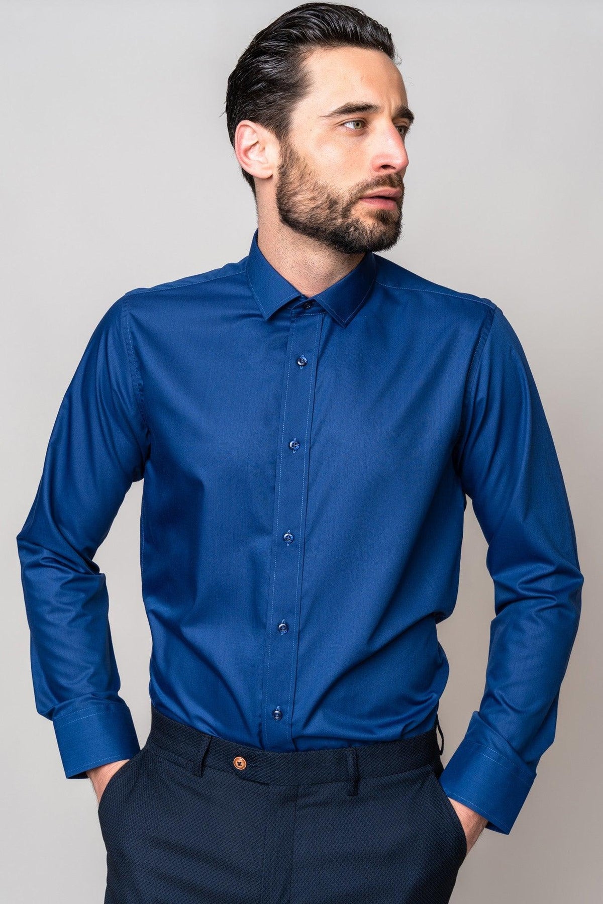 A man with dark hair and a beard stands with his hands in his pockets, wearing the Marc Darcy CARTER Navy Long Sleeve Shirt and dark trousers, looking to his left against a plain light background.