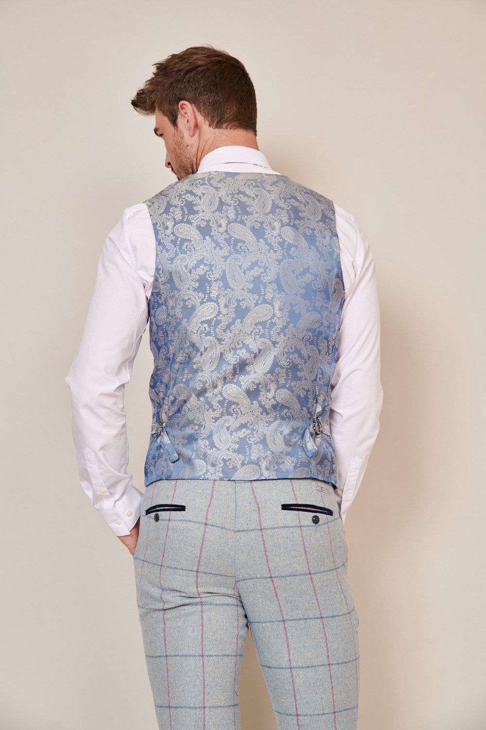 A man stands facing away, wearing the Marc Darcy NICOLAS Sky Tweed Check Waistcoat, a white dress shirt, and light gray plaid trousers—an elegant choice for a groom's suit or sky blue check occasion.