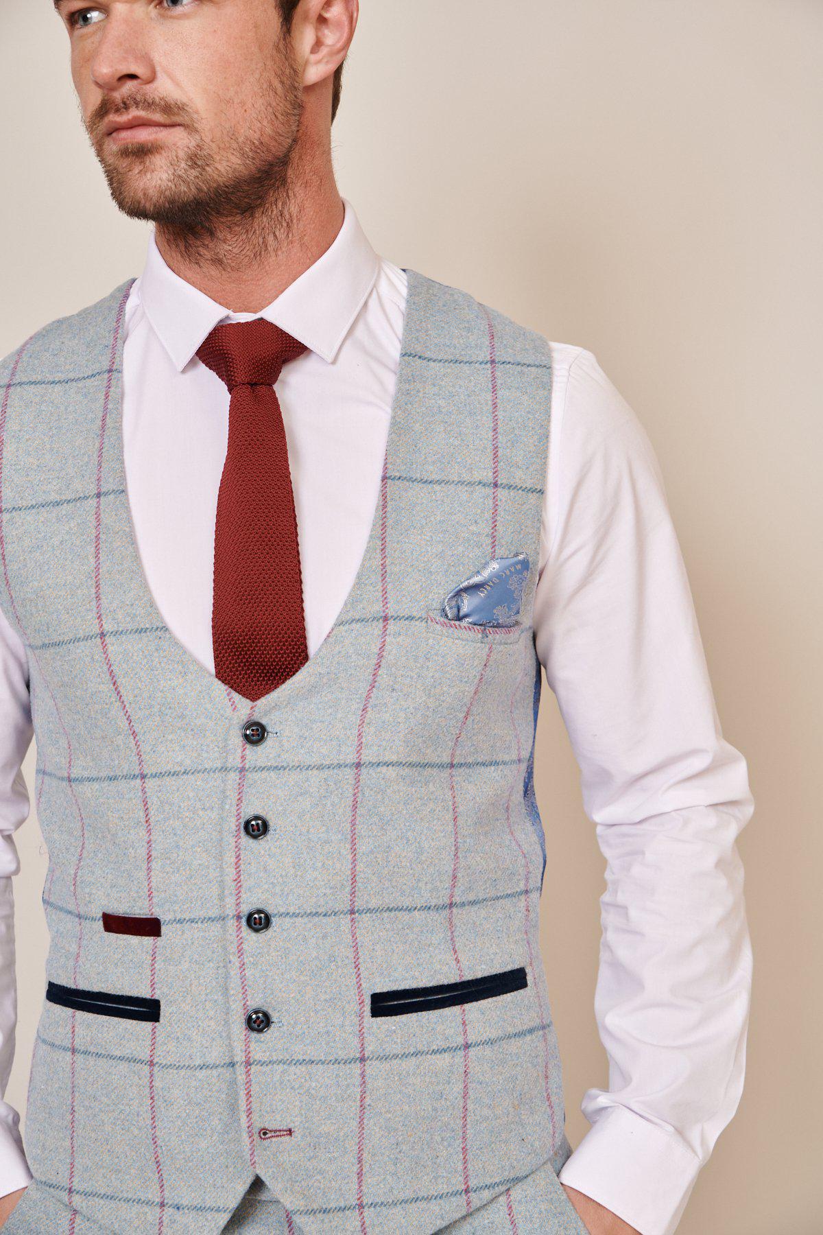 A man wears the Marc Darcy NICOLAS Sky Tweed Check Waistcoat in light gray plaid with blue and pink lines over a white shirt, red tie, and blue pocket square. The image is a chest-up shot of him looking to the side.