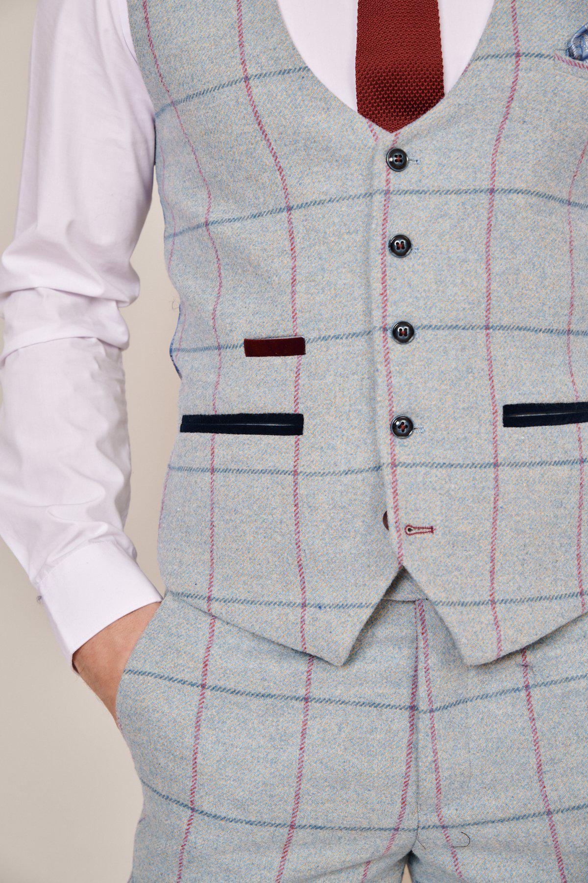A person wearing a light blue plaid suit with red and navy accents, a Marc Darcy NICOLAS - Sky Tweed Check Waistcoat, white dress shirt, and dark red tie poses with one hand in their pocket.
