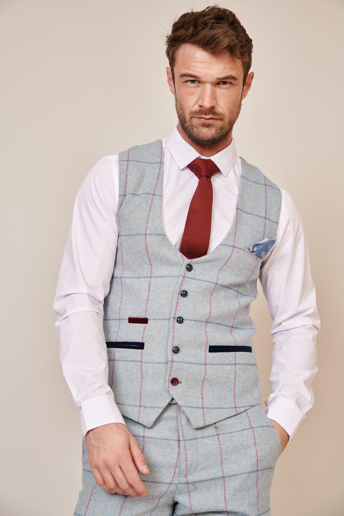 A man wearing the Marc Darcy NICOLAS Sky Tweed Check Waistcoat with matching trousers, a white dress shirt, and a red tie stands facing the camera against a plain background with one hand in his pocket.