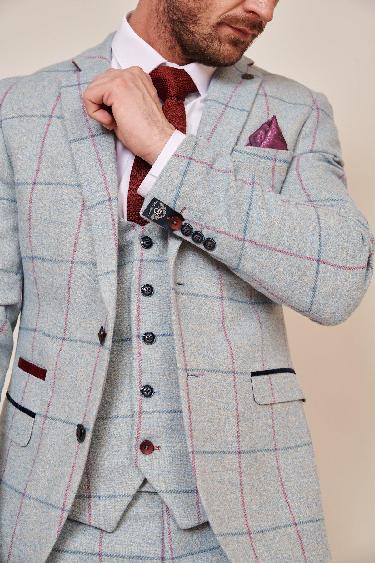 A man adjusts his tie, wearing the Marc Darcy NICOLAS - Sky Check Tweed Blazer as part of a three-piece grooms suit, paired with a burgundy knit tie, pink pocket square, and white shirt with dark buttons.