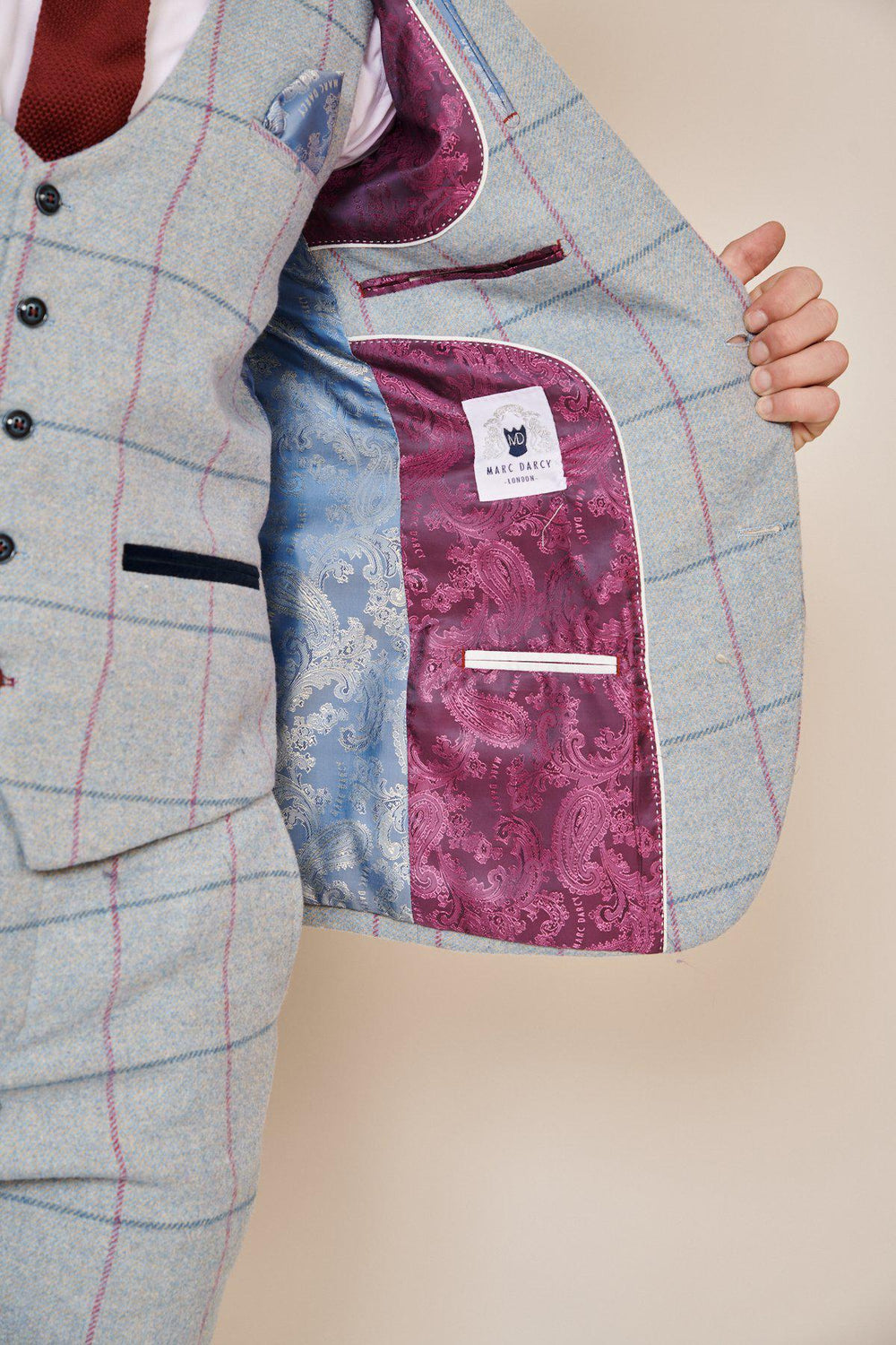 A person in a NICOLAS - Sky Check Tweed Blazer by Marc Darcy holds open the jacket, revealing a blue and burgundy paisley lining with the brand's label. Featuring black buttons and a red tie, it’s perfect for a stylish groom’s suit.