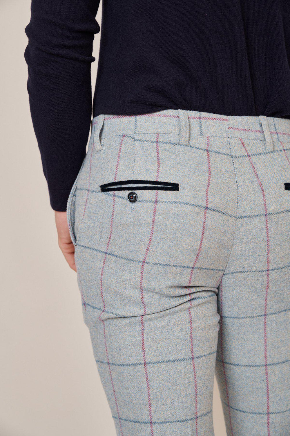 A person wears Marc Darcy NICOLAS - Sky Check Tweed Trousers, slim fit with a light gray plaid pattern featuring pink and blue lines and black buttoned back pockets; shown waist down with a dark top tucked in.