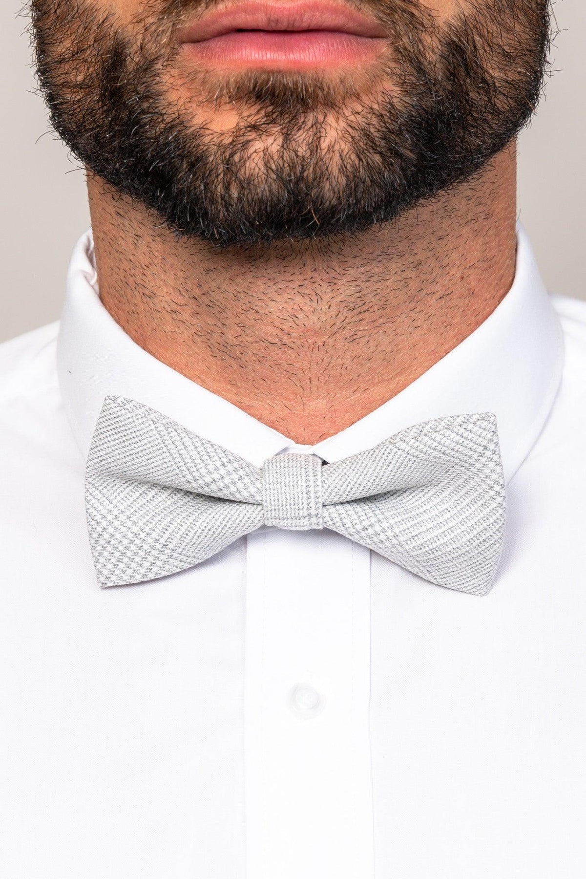 Close-up of a man in formalwear, showcasing the Accessory BROMLEY - Stone Check Bow Tie and a crisp white dress shirt. Only his neatly trimmed beard, lower face, neck, and upper chest are visible.