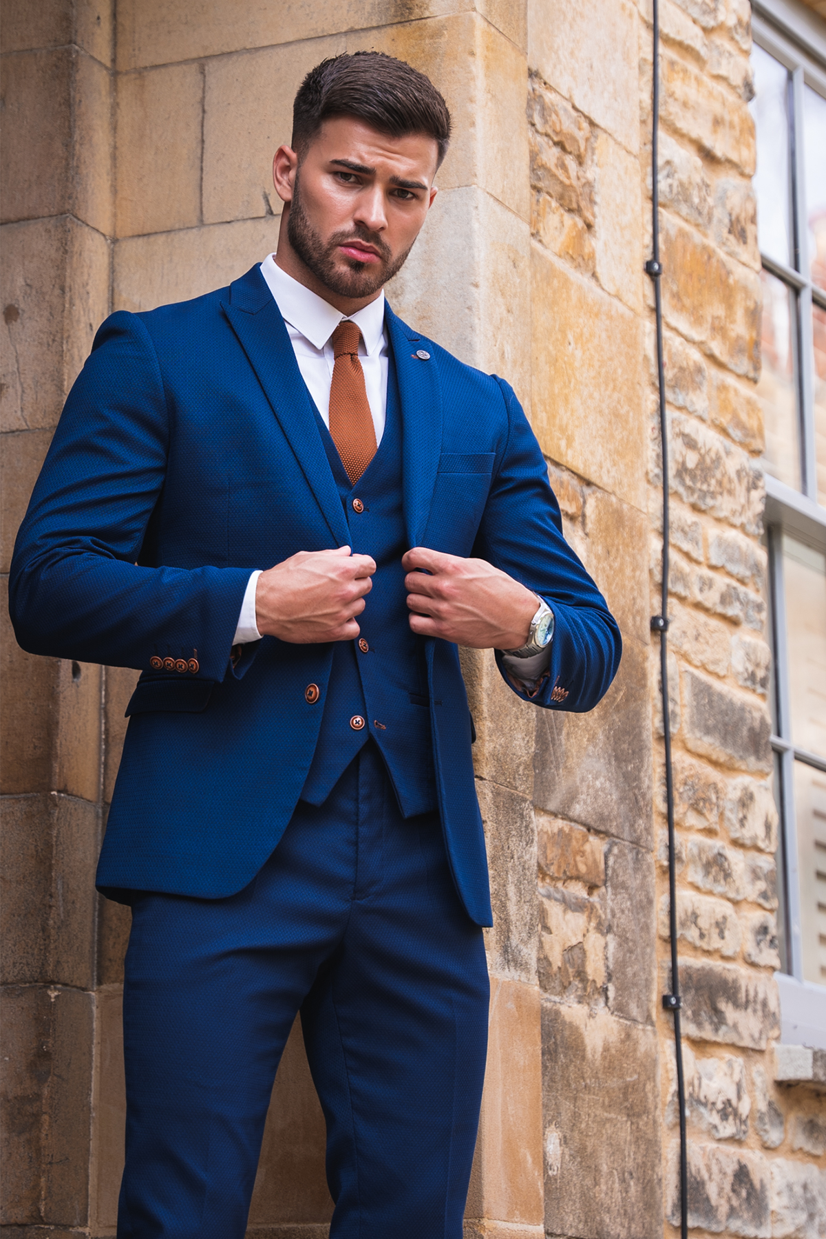 Owen Warner, runner up of 'I'm A Celebrity Get Me Out Of Here' 2022, wears a Max Royal Blue Suit by marcdarcy. He stands confidently outside near a stone wall in a white shirt, brown tie, and camouflage print pocket square.