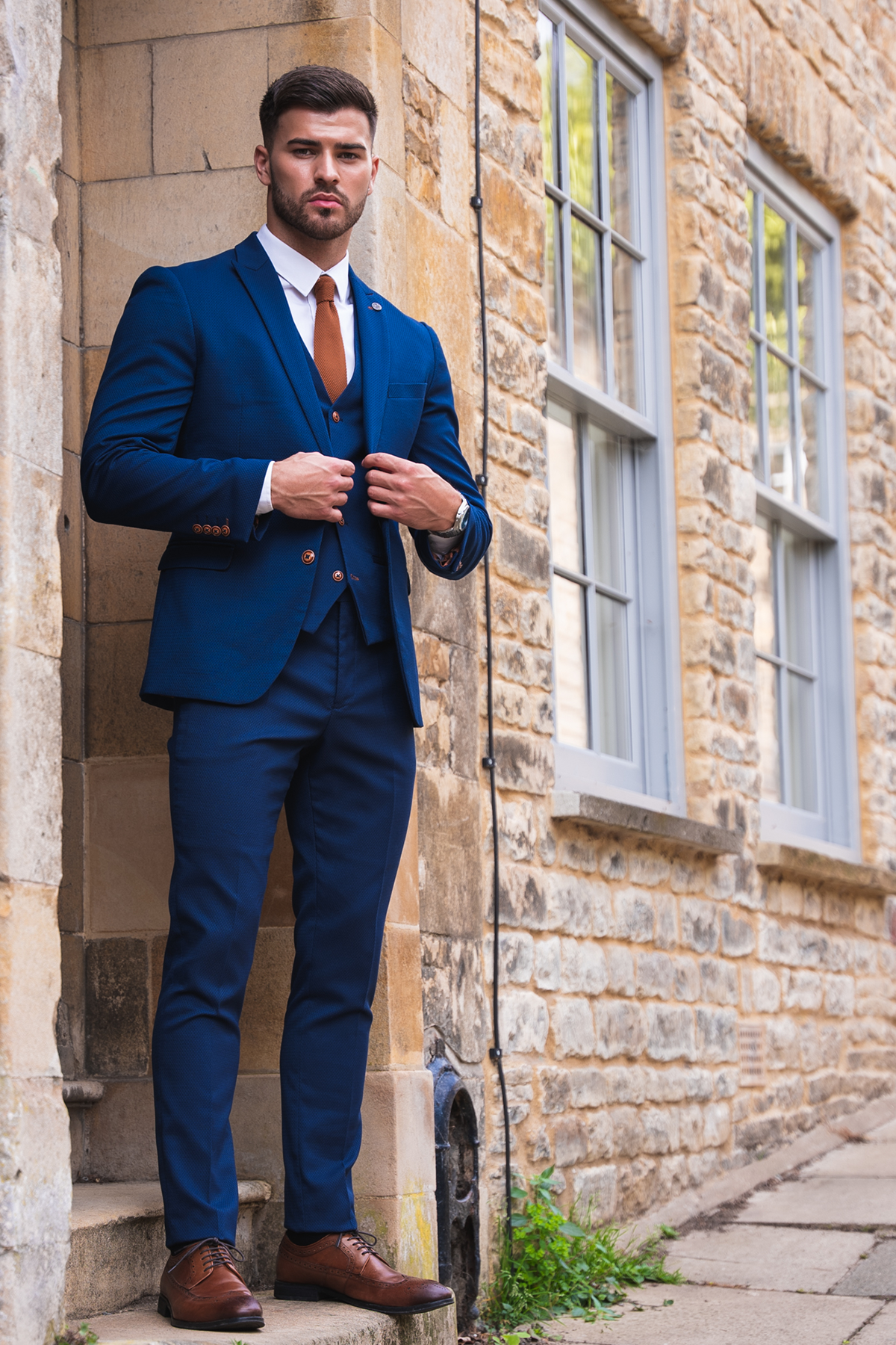 Owen Warner, 2022 'I'm A Celebrity Get Me Out Of Here' runner up, stands confidently against a stone building in a marcdarcy Max Royal Blue Suit with slim fit blazer, white shirt, rust tie, and camouflage pocket square.