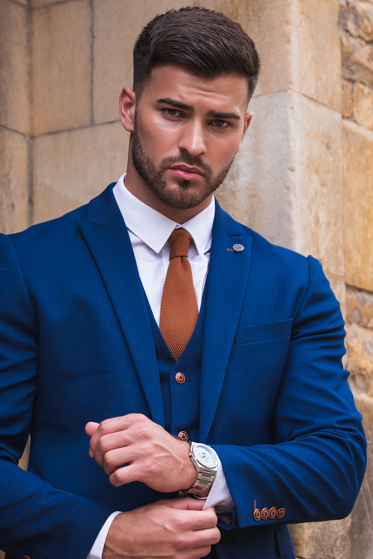 Owen Warner, 2022 'I'm A Celebrity Get Me Out Of Here' runner up, poses confidently in a marcdarcy Max Royal Blue Suit with a slim fit blazer and camouflage pocket square, adjusting his silver watch against a stone wall.