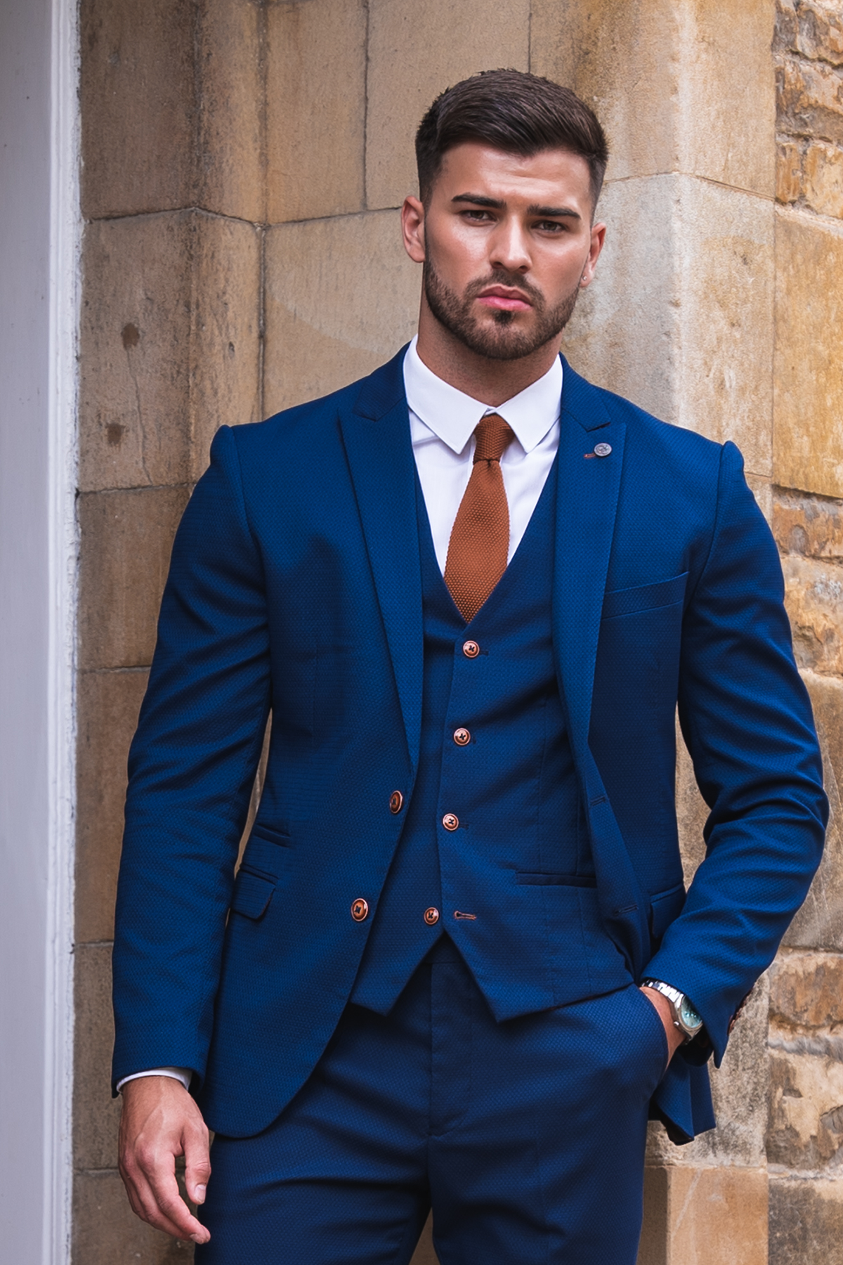 Owen Warner, 2022 'I'm A Celebrity Get Me Out Of Here' runner-up, wears a marcdarcy Max Royal Blue Suit with a slim-fit blazer, white shirt, rust tie, and camouflage pocket square as he leans against a stone wall.