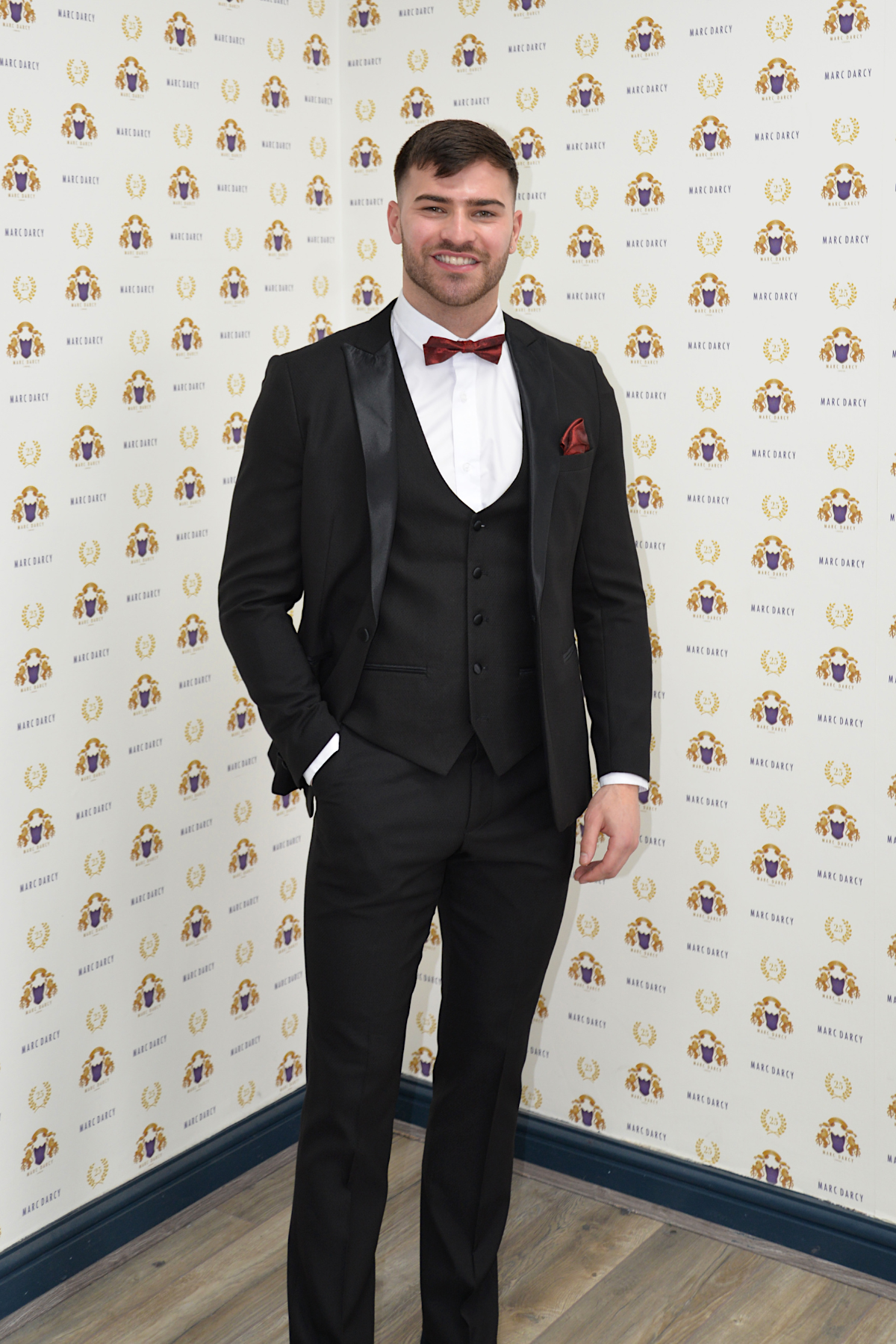 Owen Warner, 2022 'I'm A Celebrity Get Me Out Of Here' runner up, smiles in a marcdarcy Dalton black tuxedo with a red bow tie and pocket square, posing before a Huge Daddy step-and-repeat featuring a colorful lion logo.