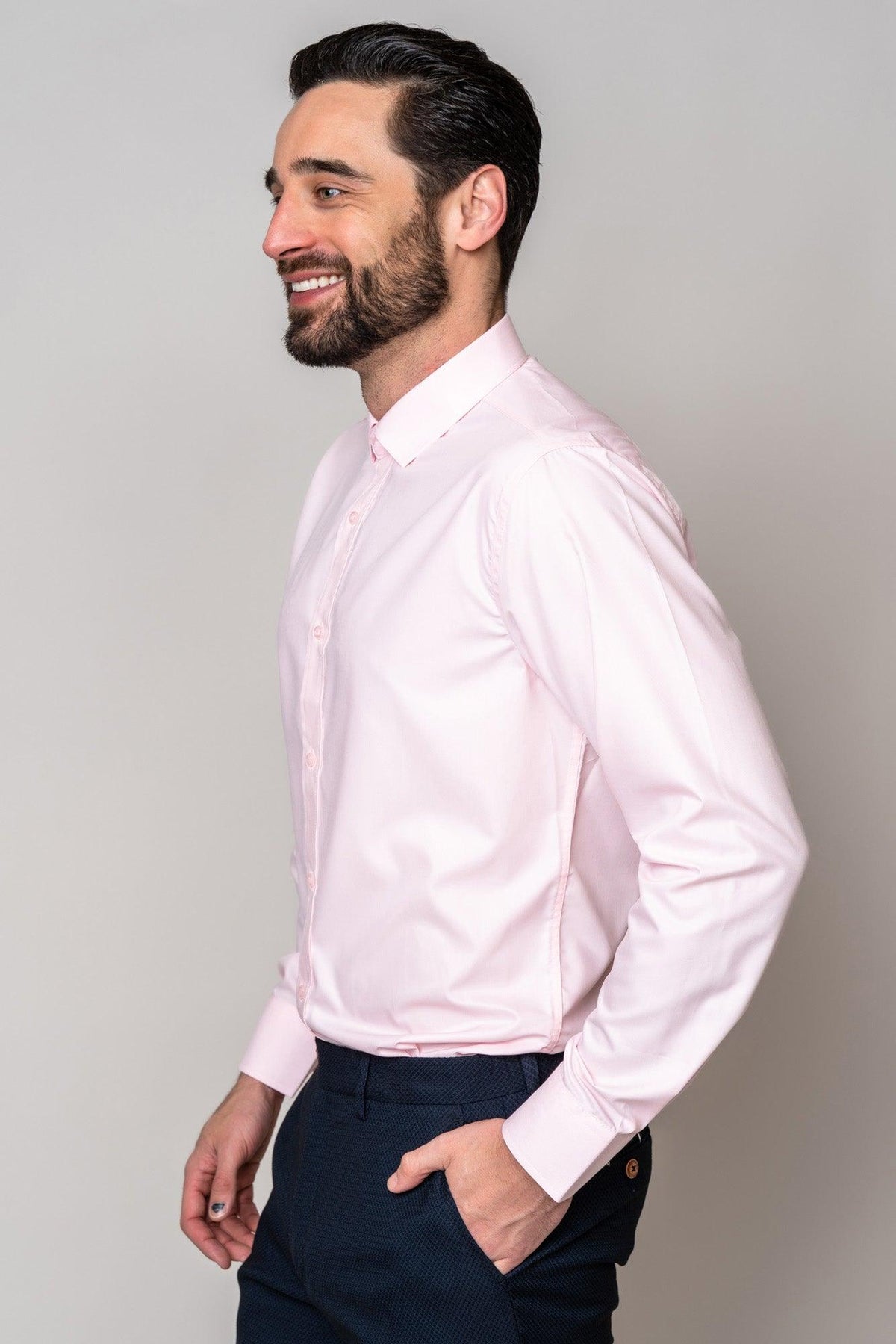 A man with dark hair and a beard smiles in profile, wearing the Marc Darcy CARTER pink long sleeve shirt and dark pants. He stands with one hand in his pocket against a plain background.