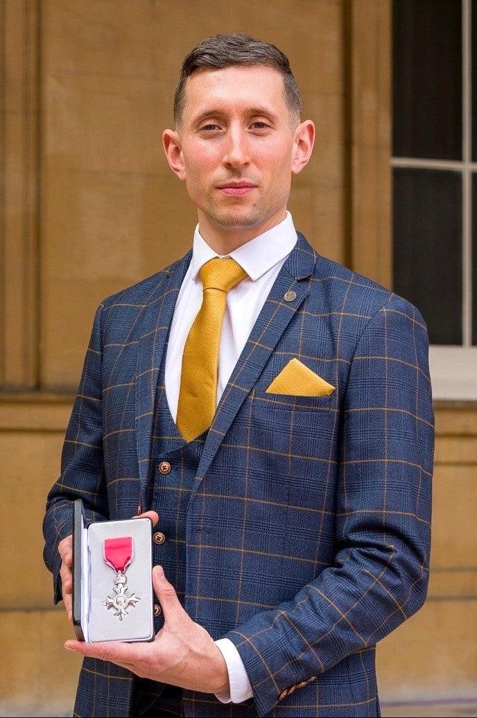 Paralympian Jonathan Broom-Edwards MBE, in a marcdarcy Jenson Marine Check Suit and yellow tie, stands outdoors before a tan building, holding an open box displaying a medal on a pink ribbon.