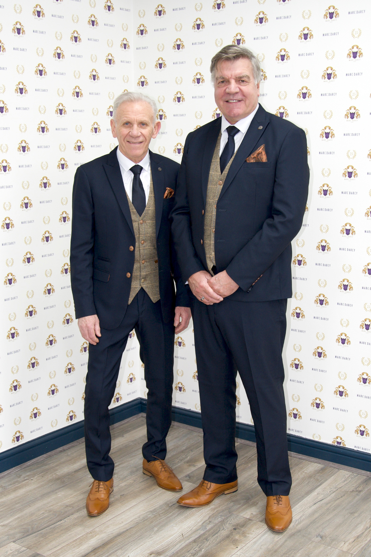 Former Everton Midfielder Peter Reid and Football Manager Sam Allardyce wear the marcdarcy Max Navy Suit with Ted Waistcoat, posing before a white wall patterned with the marcdarcy logo.